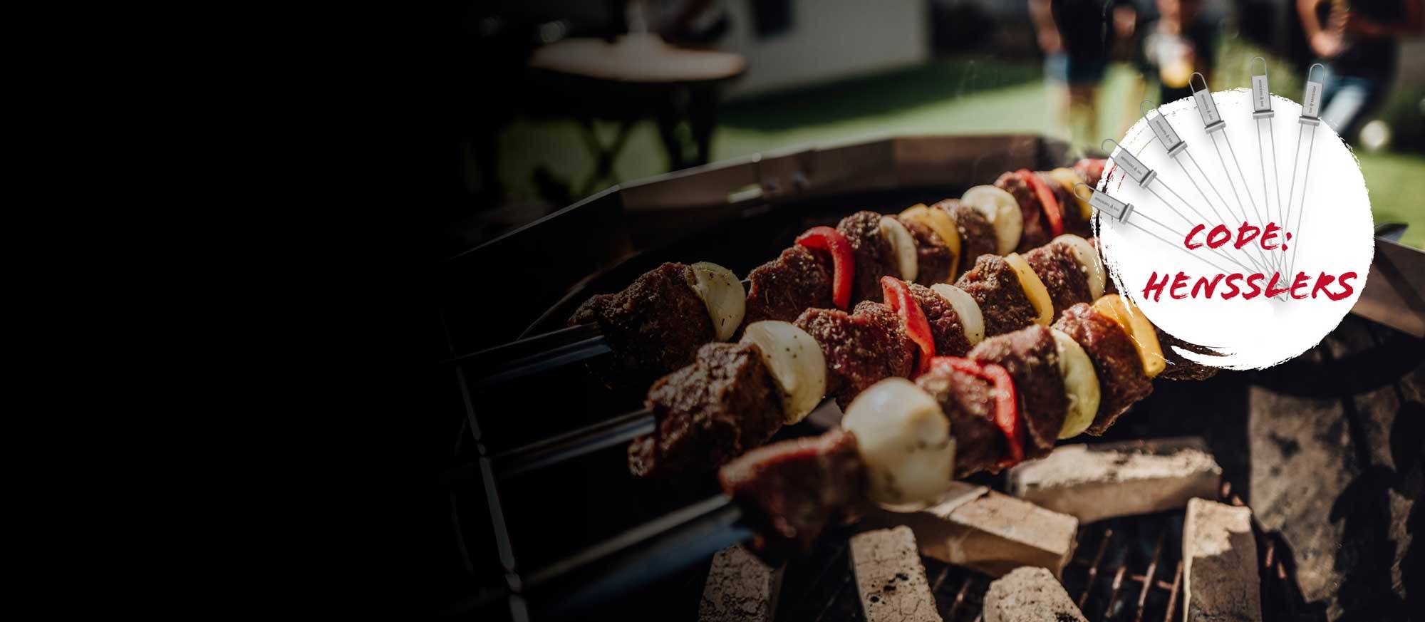 Nahaufnahme von mehreren Fleisch- und Gemüsespießen (Schaschlik) mit Zwiebeln und Paprika auf einem Grill. Im oberen rechten Bereich ein weißer Kreis mit rotem Text "CODE: HENSSLERS" und mehreren Metall-Grillspießen.