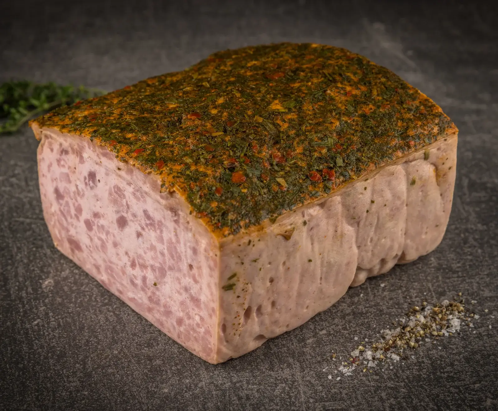 Ein großer, rechteckiger Block Fleischkäse, oben reichlich mit grünen Kräutern und roten Gewürzen bestreut. Eine Schnittfläche zeigt die hellrosa, feine Textur. Auf dunklem Untergrund mit Salz und Pfeffer.