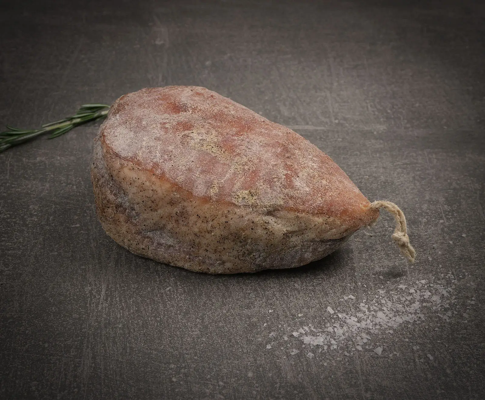 Ein Stück luftgetrocknete Wurst mit weißem Edelschimmel und Pfefferkörnern liegt auf einem dunklen, strukturierten Untergrund. Links ist ein Rosmarinzweig, rechts grobe Meersalzflocken zu sehen. Am Ende ist ein Bindfaden.