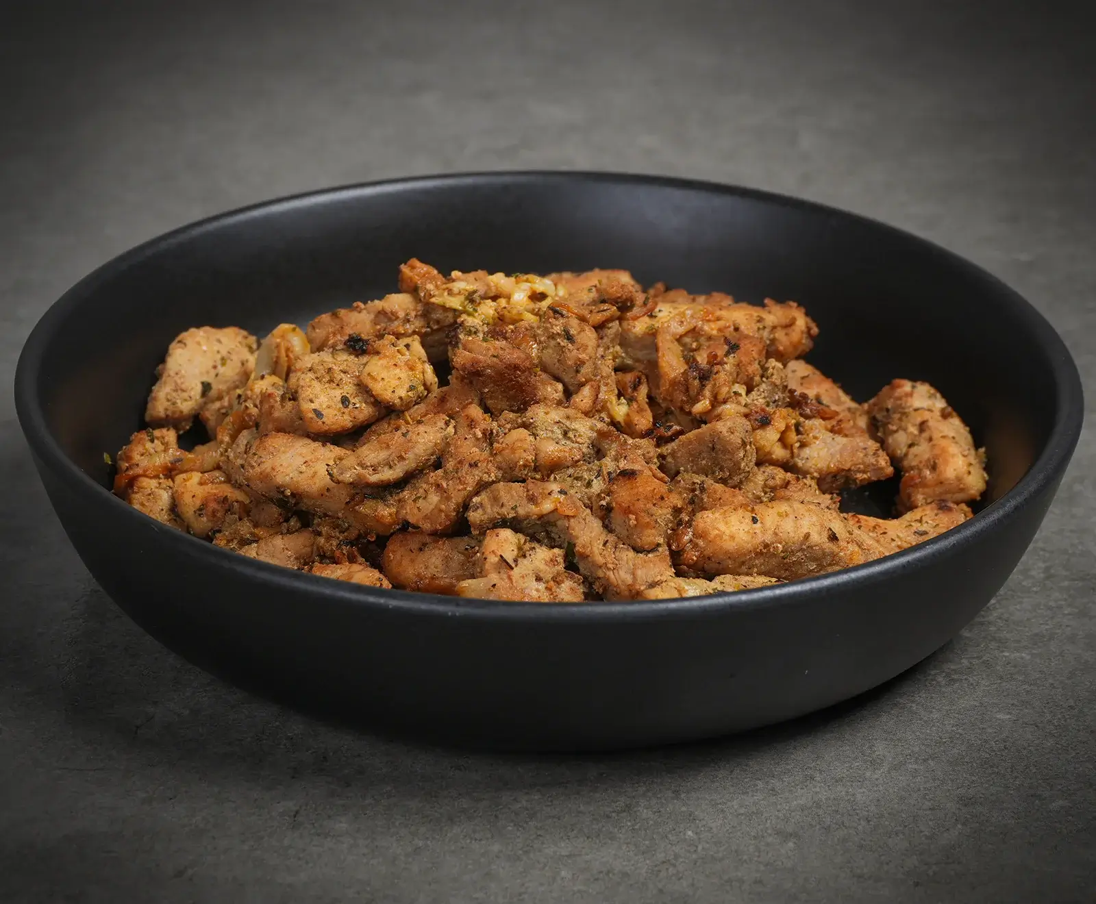 Eine schwarze Schale gefüllt mit zahlreichen goldbraun gebratenen und gewürzten Fleischstücken, präsentiert auf einem dunklen, grauen Untergrund.