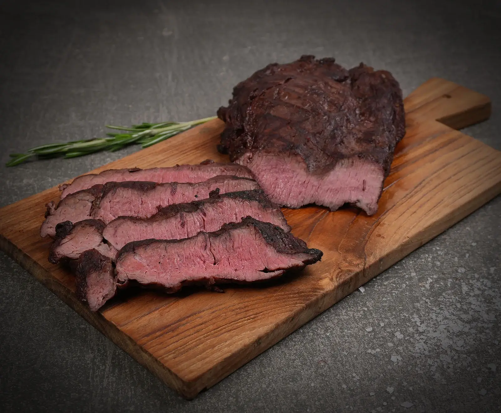 Ein aufgeschnittenes, medium-rosa gebratenes Stück Fleisch auf einem Holzbrett, mit mehreren Scheiben daneben und einem Rosmarinzweig im Hintergrund. Der dunkle Untergrund ist leicht mit Salzkristallen bestreut.