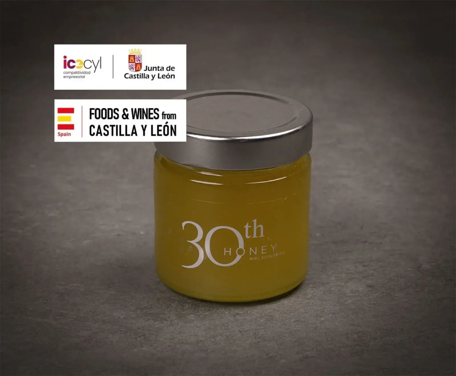 Ein Glas mit gelbem Honig und silbernem Deckel, beschriftet mit "30th HONEY Miel Ecológica". Oben links Logos von "Junta de Castilla y León" und "Foods & Wines from Castilla y León".