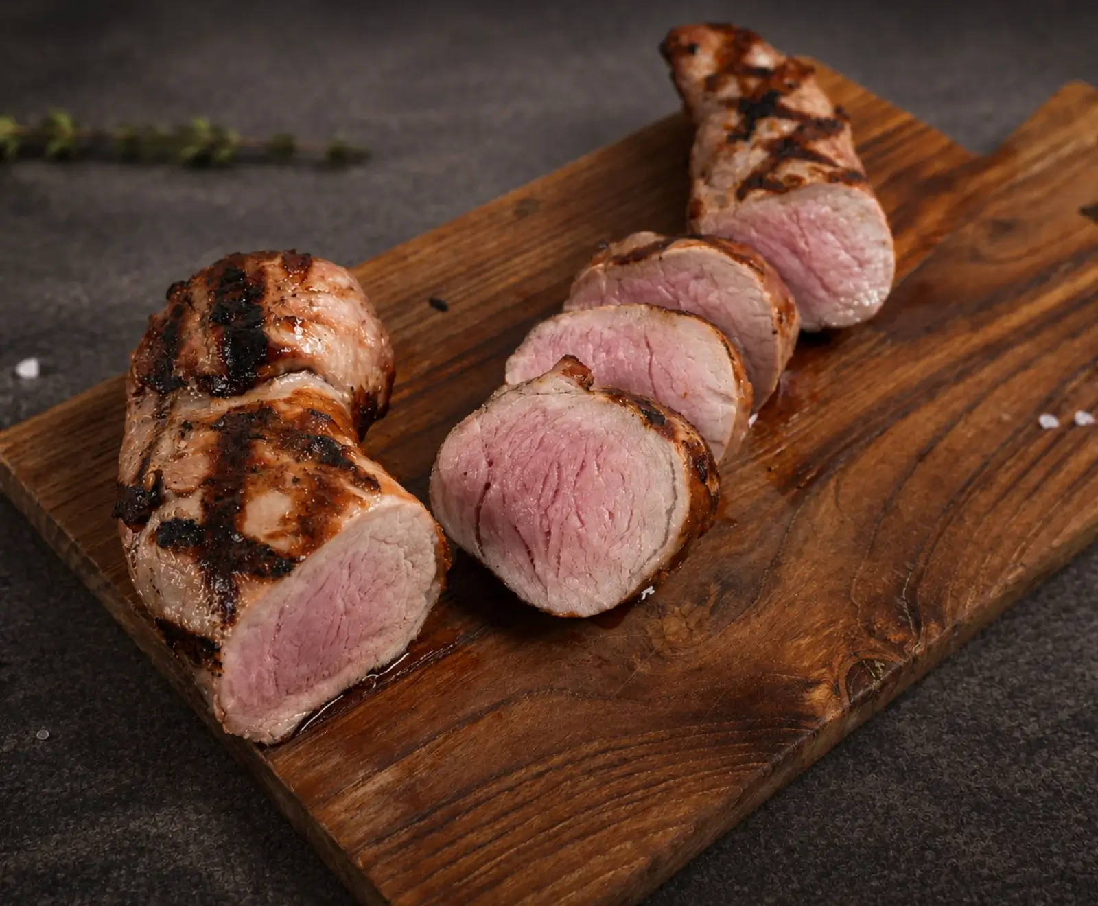 Gegrilltes Schweinefilet auf einem Holzbrett, teilweise in dicke Scheiben geschnitten mit rosa Kern und deutlichen Grillspuren. Grobes Salz und ein Kräuterzweig im Hintergrund.