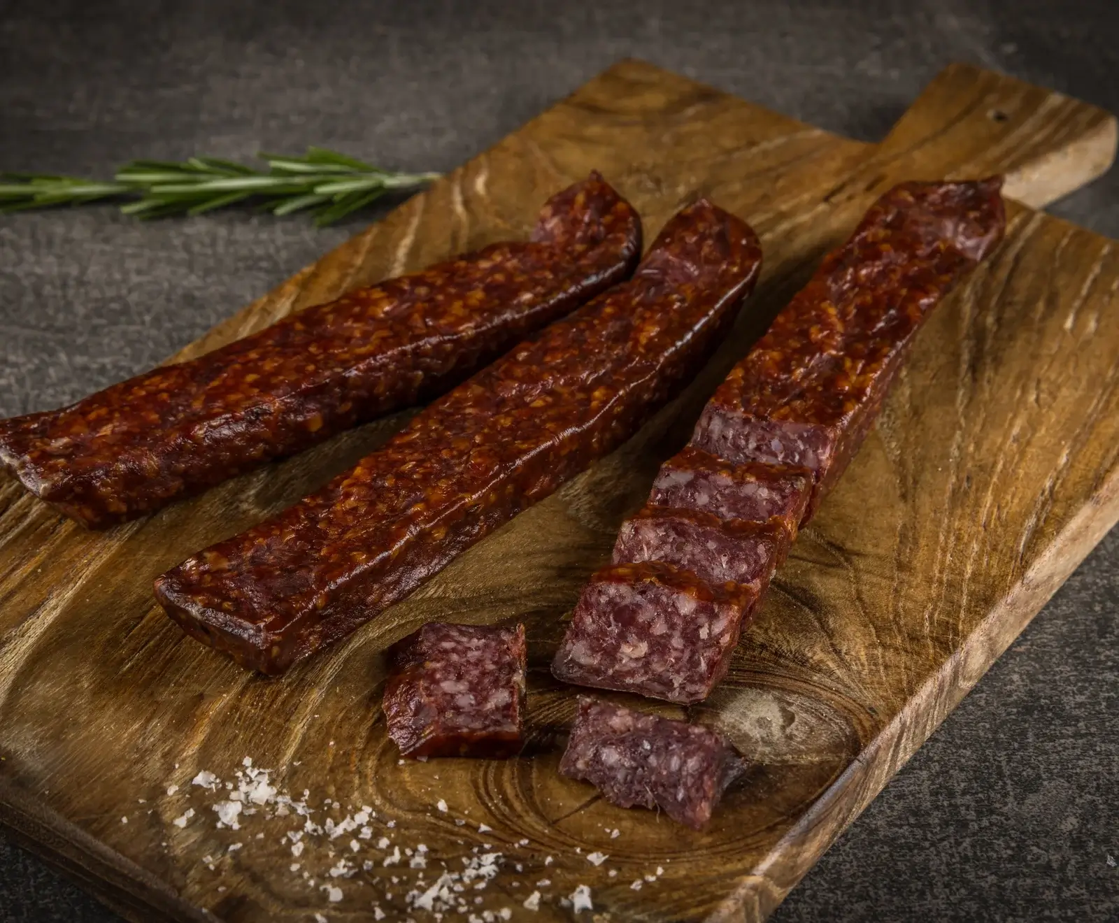 Drei dunkelrote, rechteckige Rohwürste auf einem Holzbrett. Eine Wurst ist in Scheiben geschnitten, daneben liegen Salzflocken und ein Rosmarinzweig. Der Hintergrund ist dunkel.