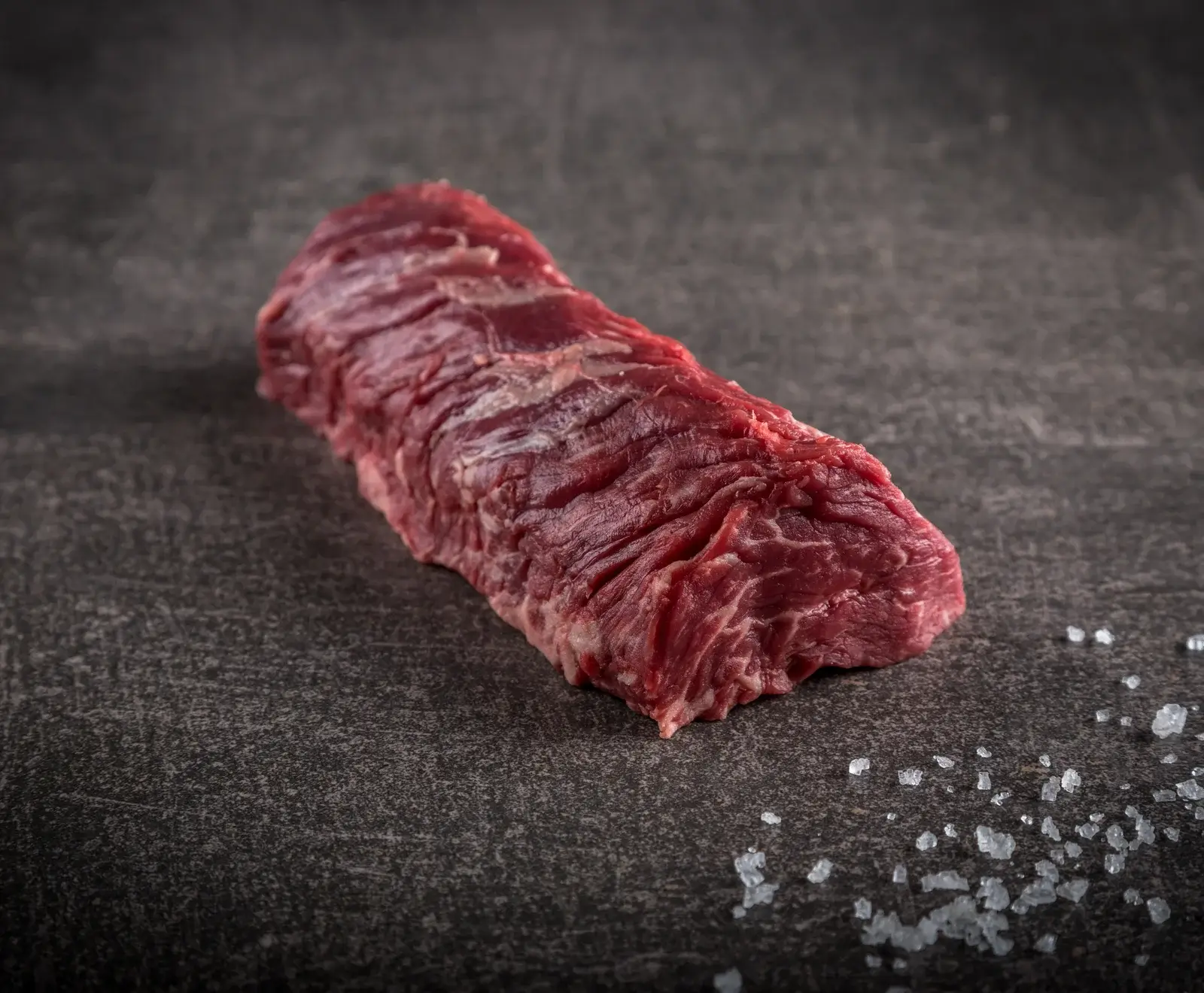 Ein rohes, längliches Stück Rindfleisch mit sichtbarer Faserung liegt auf einer dunklen, strukturierten Oberfläche. Grobe Meersalzkristalle sind daneben verstreut.
