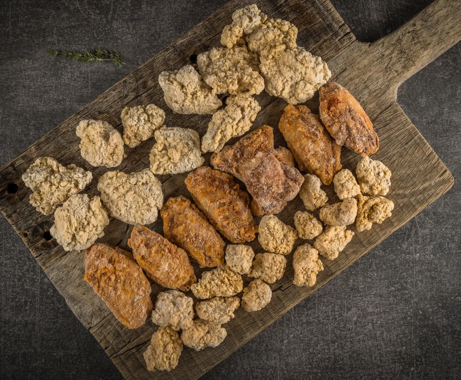 Draufsicht auf eine rustikale Holzplatte mit gefrorenen, panierten Hähnchenflügeln (rötlich-braun) und Hähnchen-Nuggets (hellbeige). Ein Thymianzweig liegt oben links auf dunklem Hintergrund.