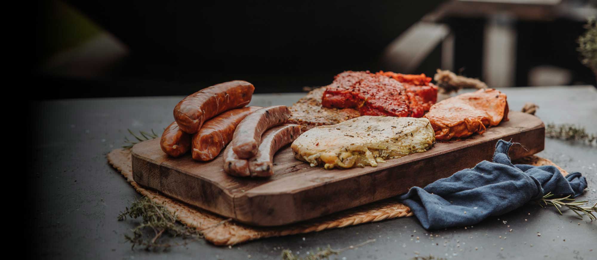 Eine Holzplatte mit verschiedenen rohen Fleischstücken, darunter Würste, mariniertes Steak und Kotelett, bereit zum Grillen. Rosmarin und Thymian liegen daneben.