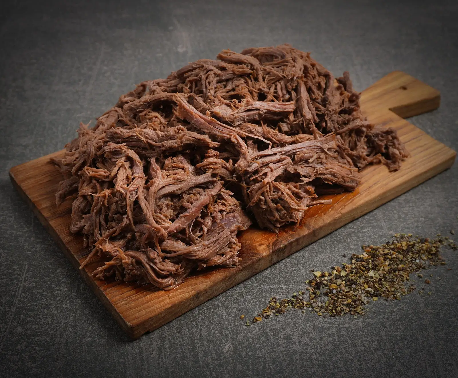 Ein großer Haufen zerkleinertes, braunes Fleisch (Pulled Pork oder Beef) auf einem Holzschneidebrett, daneben eine Prise grober Pfeffer auf dunklem Untergrund.