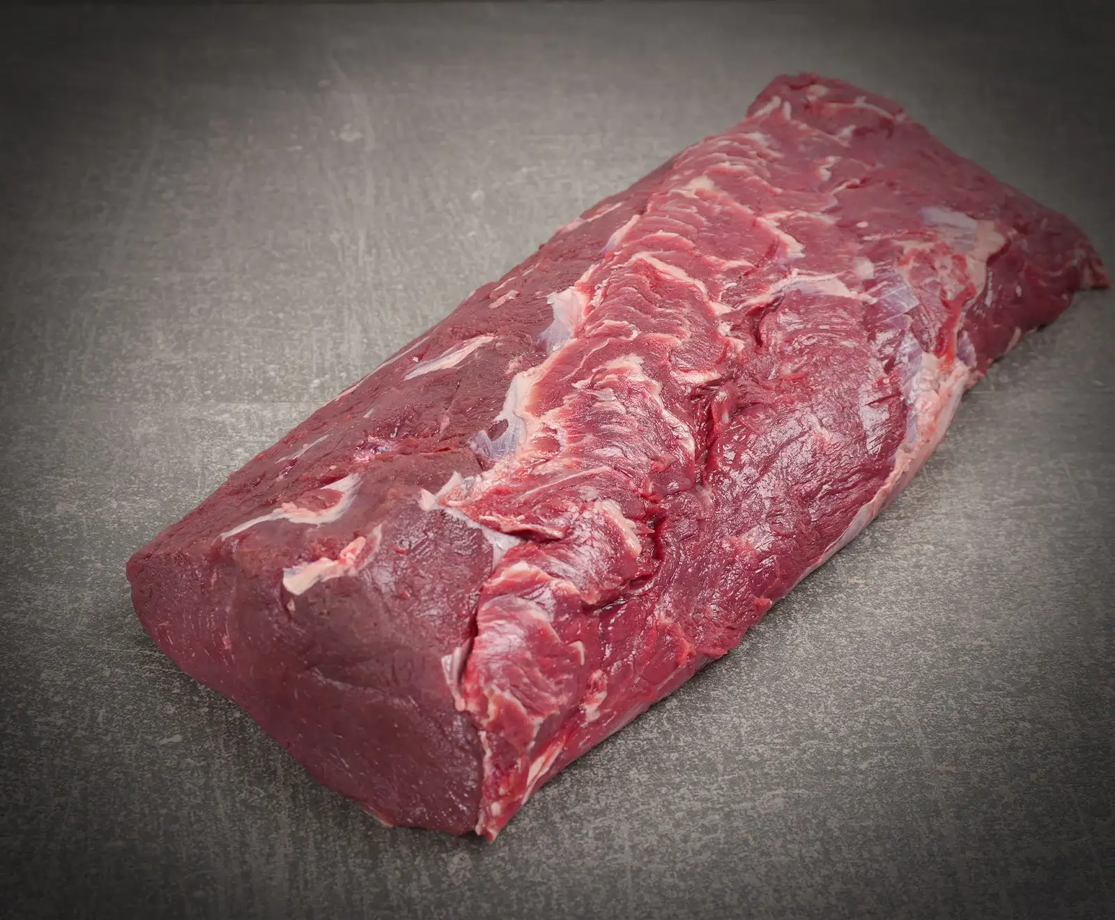 Ein großes, rohes Stück tiefrotes Rindfleisch mit sichtbaren Fettadern liegt auf einer dunklen, strukturierten Oberfläche. Es ist frisch und bereit zur Zubereitung.
