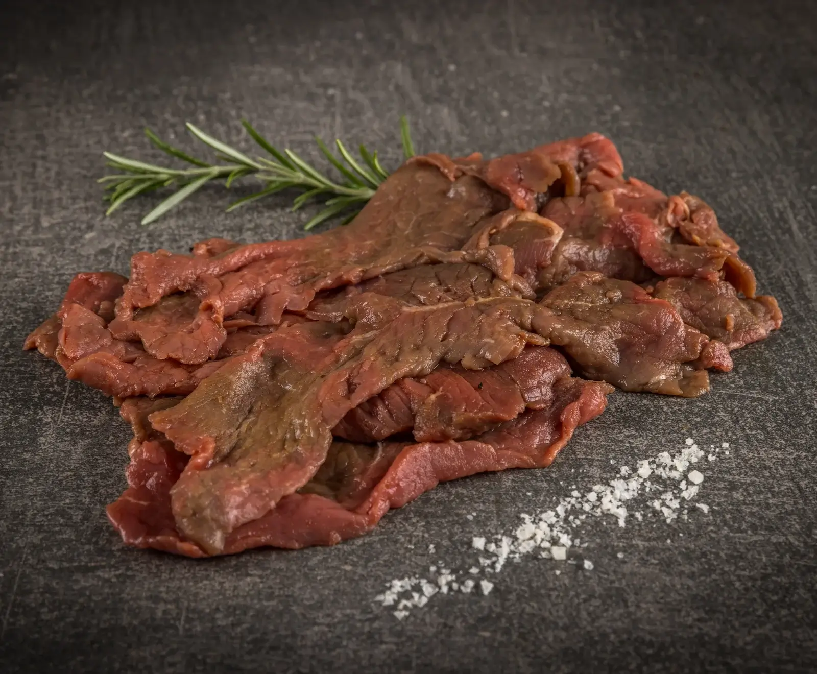 Dünn geschnittenes rohes Rindfleisch auf dunklem Untergrund, garniert mit einem Rosmarinzweig und grobem Salz.