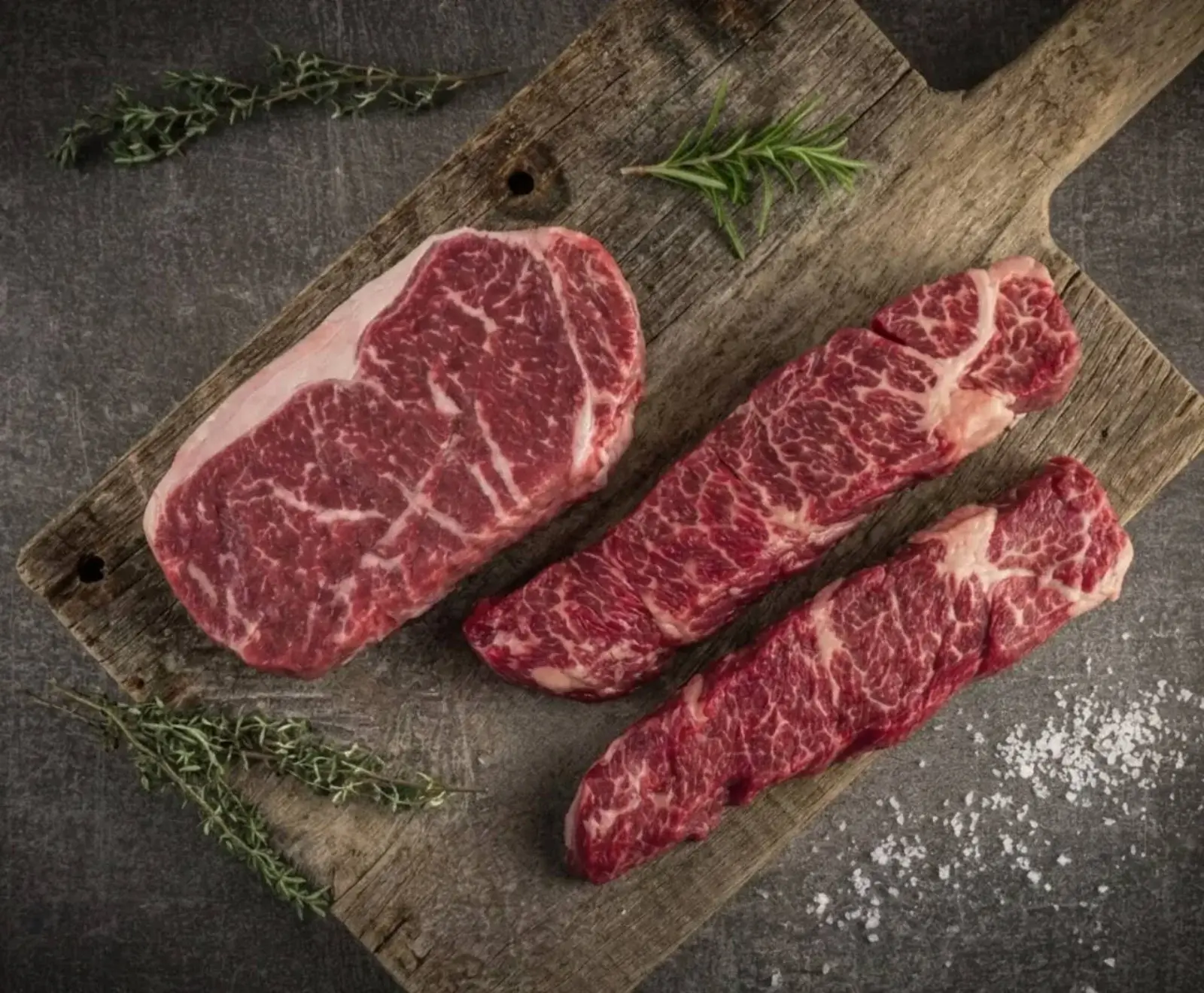 Drei rohe Rindersteaks mit deutlicher Marmorierung liegen auf einem rustikalen Holzbrett. Rosmarin, Thymian und grobes Salz sind um die Steaks auf dunklem Untergrund verteilt.