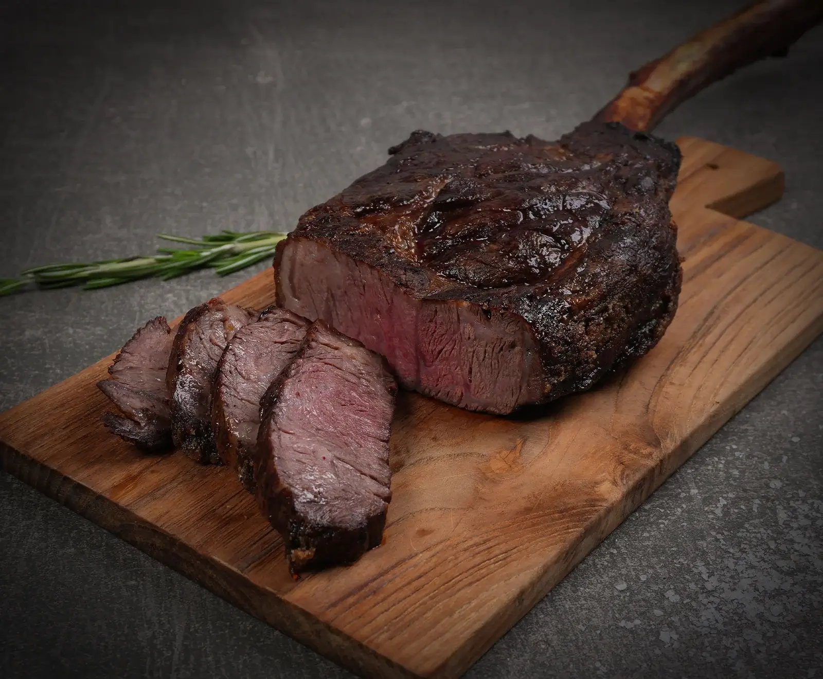 Ein perfekt gegartes Tomahawk-Steak, teilweise in Scheiben geschnitten, auf einem Holzschneidebrett. Das Fleisch ist innen rosa und außen knusprig. Ein Rosmarinzweig liegt daneben.