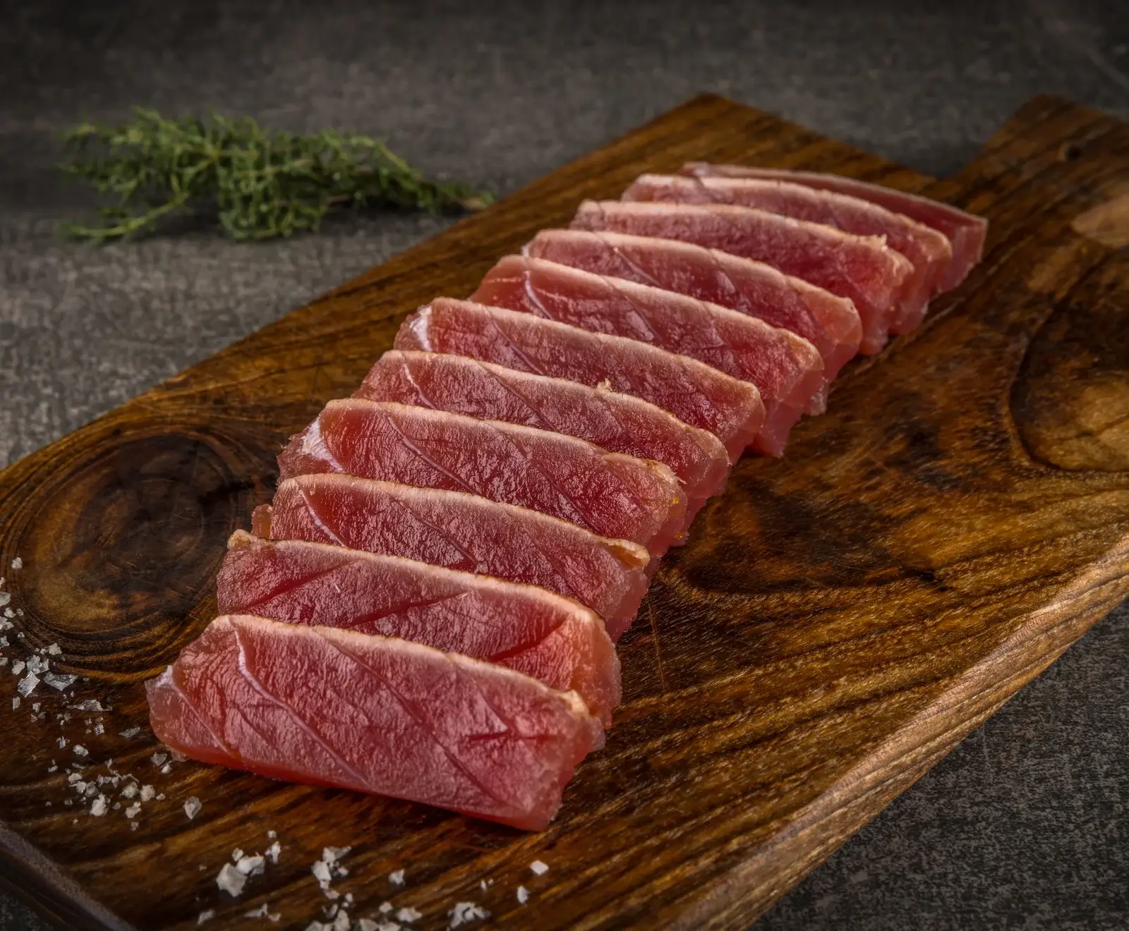 Dünne Scheiben rohen, tiefroten Thunfischs liegen schräg auf einem dunklen Holzbrett, bestreut mit Meersalz. Im Hintergrund ist ein Kräuterzweig auf dunklem Untergrund zu sehen.