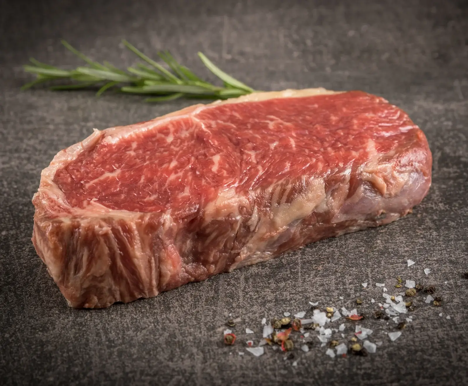 Ein rohes, marmoriertes Rindersteak liegt auf einer dunklen, strukturierten Oberfläche. Daneben sind ein Rosmarinzweig, grobes Salz und bunte Pfefferkörner zu sehen.