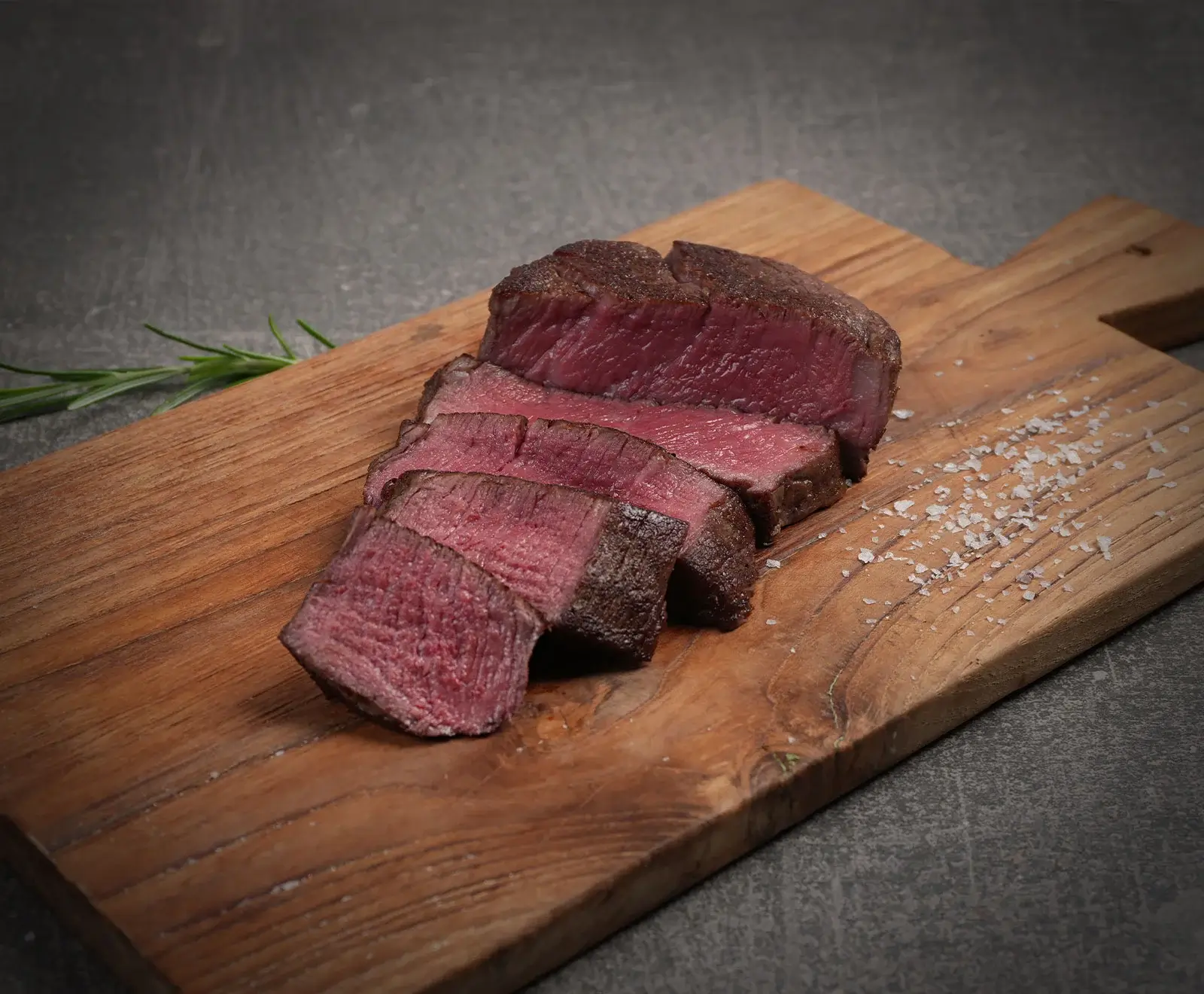 Ein perfekt gegartes, in Scheiben geschnittenes Steak (medium-rare) liegt auf einem Holzschneidebrett. Grobe Salzkristalle und ein Rosmarinzweig sind daneben zu sehen, auf dunklem Hintergrund.