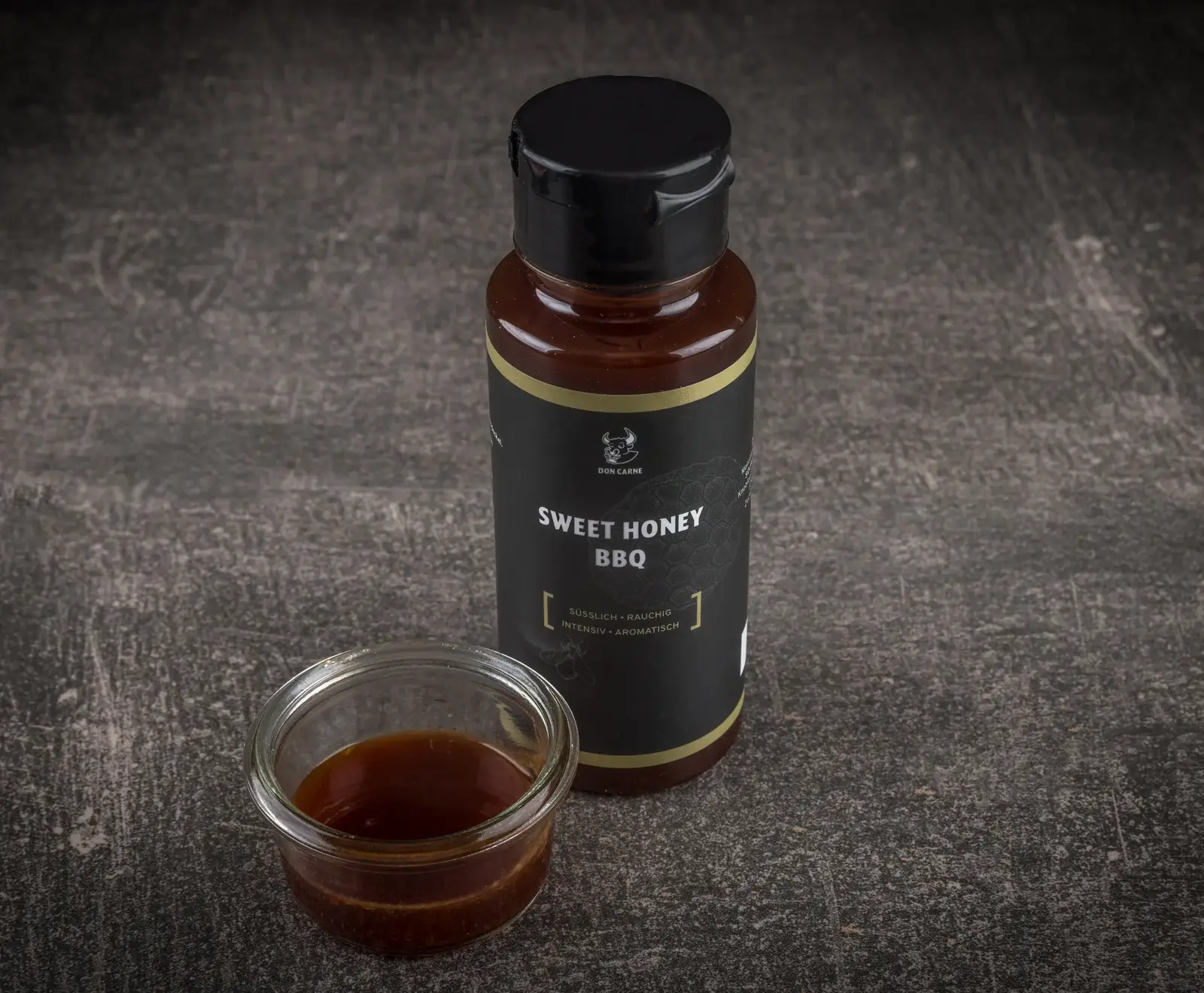 Eine dunkle Flasche Don Carne Sweet Honey BBQ Sauce mit schwarzem Etikett und goldenen Akzenten steht auf einem dunklen Untergrund. Daneben ist ein kleines Glas mit der braunen Sauce zu sehen.