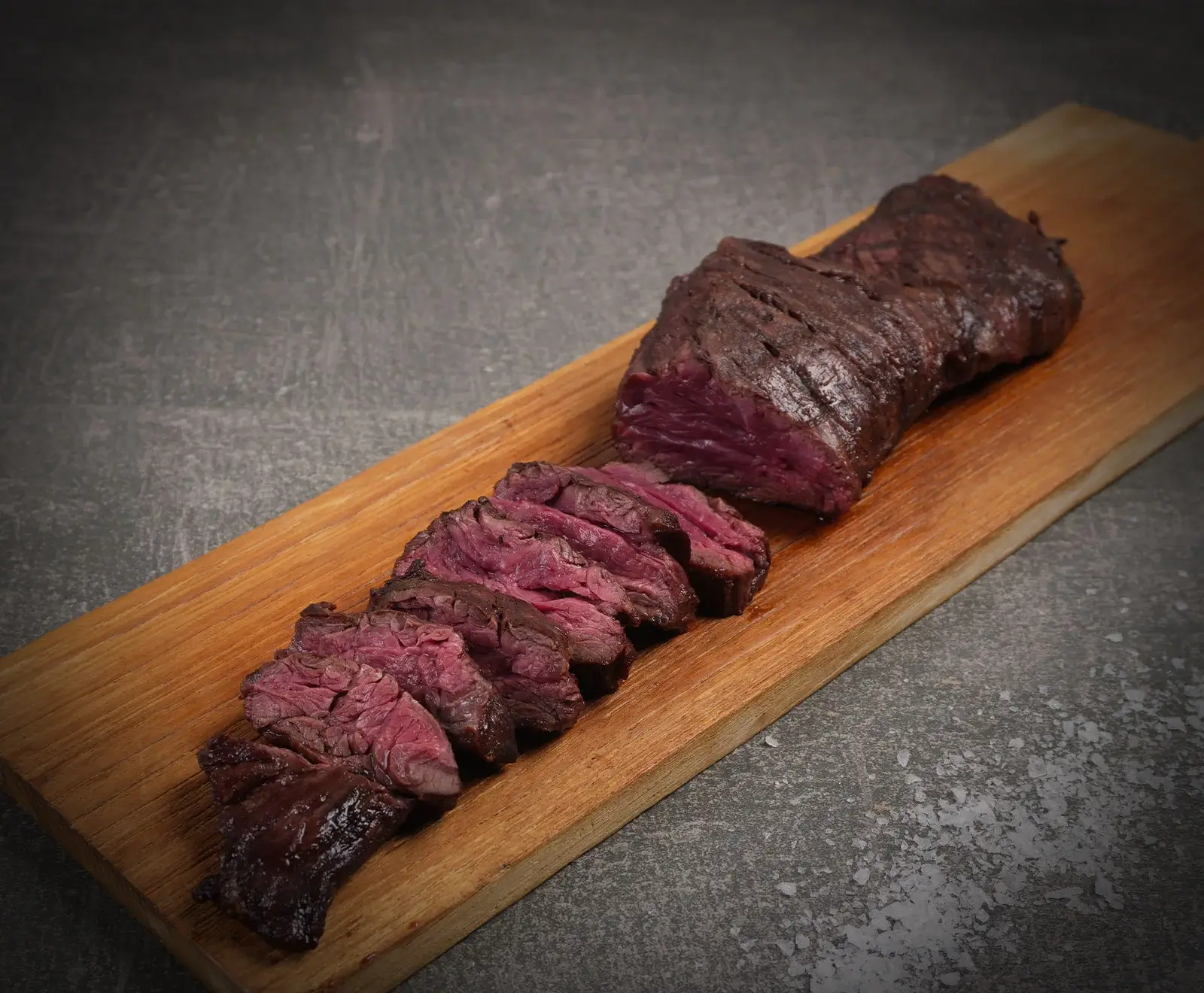 Geschnittenes, medium-rare gebratenes Fleischstück mit dunkler Kruste und rotem Kern auf einem Holzbrett. Der Hintergrund ist dunkelgrau mit groben Salzkristallen.