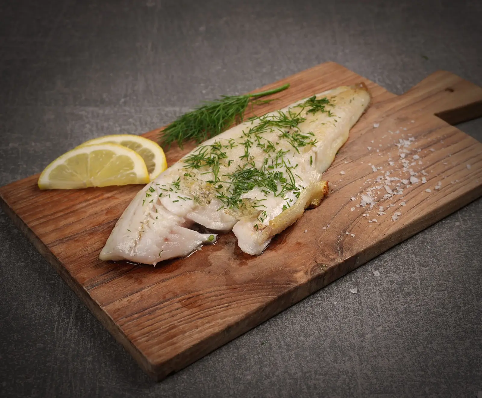 Ein gebratenes Fischfilet mit frischem Dill, zwei Zitronenspalten und grobem Meersalz auf einem rustikalen Holzbrett vor dunklem Hintergrund.
