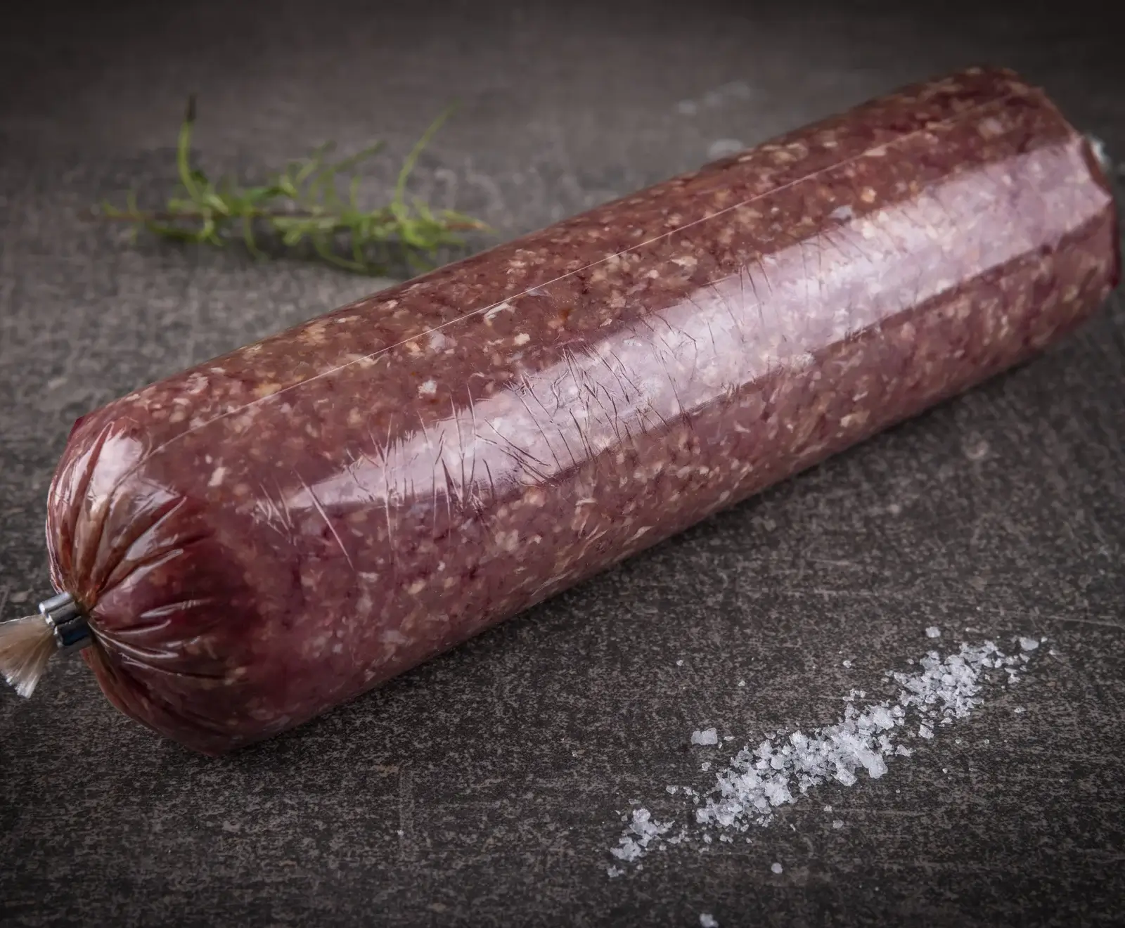 Eine längliche, in Klarsichtfolie verpackte Rohwurst (Mettwurst oder Teewurst) liegt diagonal auf einem dunklen, strukturierten Untergrund. Im Hintergrund ist ein Rosmarinzweig, im Vordergrund grobes Salz.