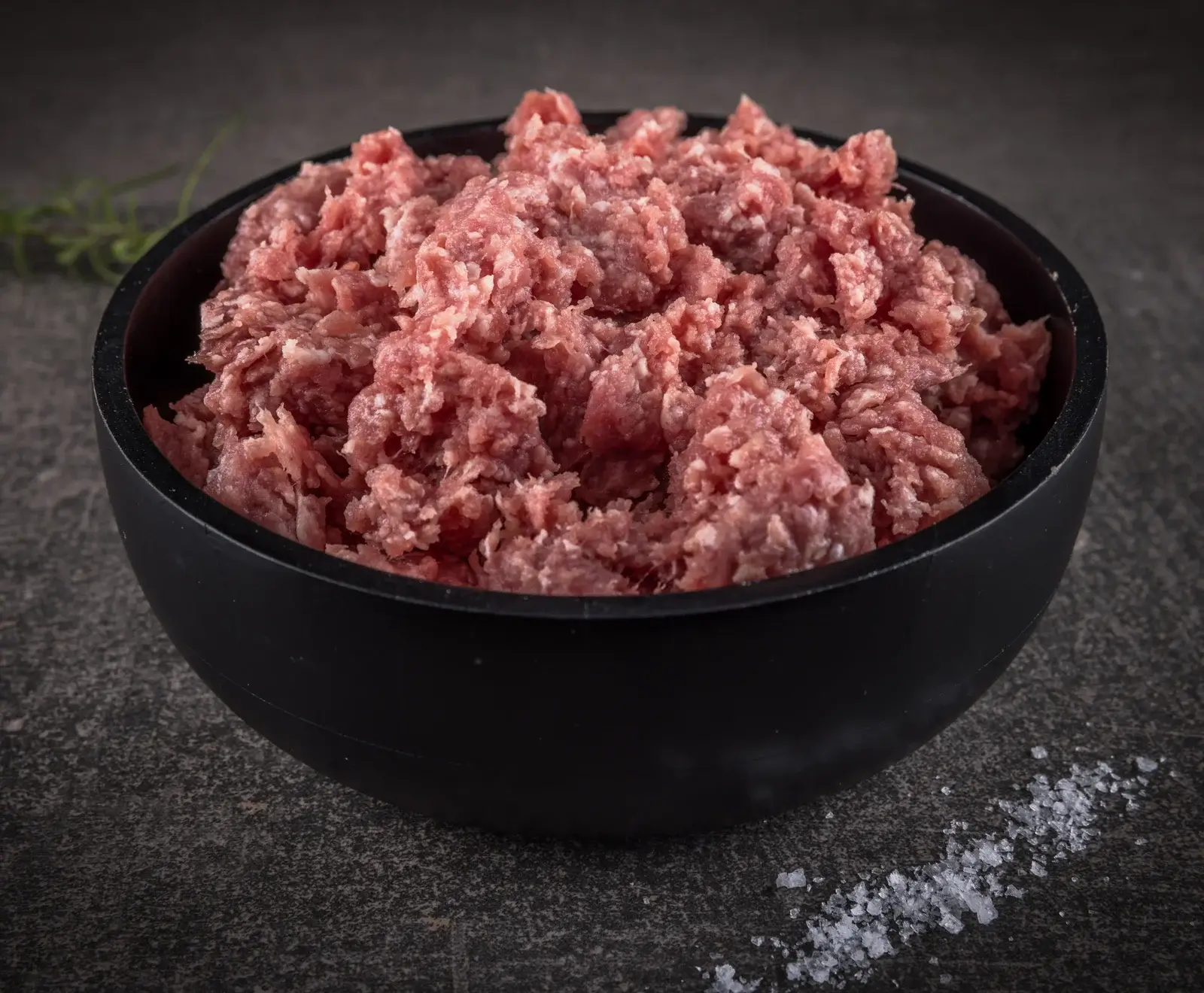 Eine schwarze Schale gefüllt mit rohem, rosa-rotem Hackfleisch, auf einer dunklen Oberfläche. Im Hintergrund ein unscharfer Rosmarinzweig, im Vordergrund grobe Salzkristalle.