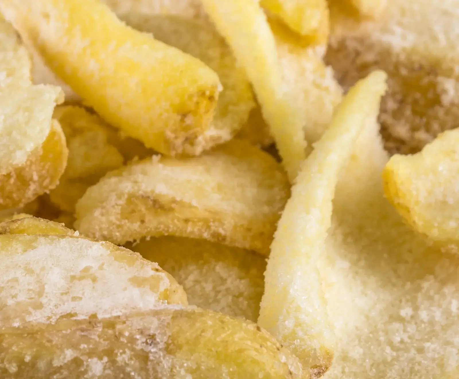 Nahaufnahme von gefrorenen Pommes Frites oder Kartoffelspalten, die mit einer Schicht aus Eiskristallen bedeckt sind. Die Kartoffelstücke sind hellgelb bis leicht goldbraun und haben unregelmäßige Formen.