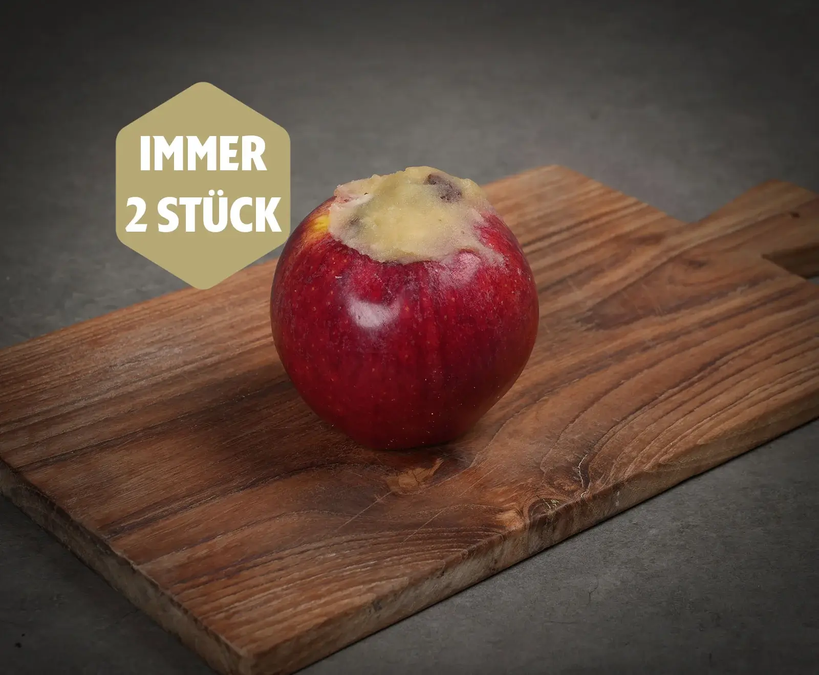 Ein roter Apfel, dessen Kern entfernt und mit einer hellen Füllung versehen wurde, liegt auf einem Holzschneidebrett. Oben links ein goldenes Etikett mit der Aufschrift "IMMER 2 STÜCK".