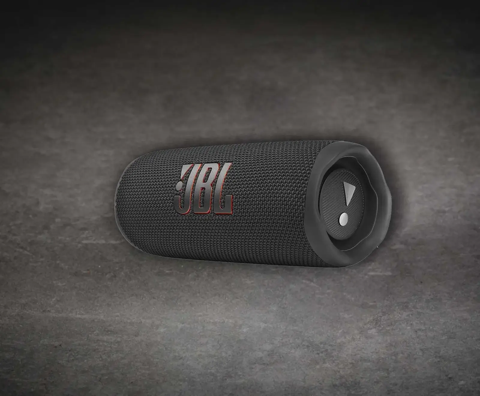 Ein schwarzer, zylindrischer JBL Bluetooth-Lautsprecher mit Stoffbezug und rotem JBL-Logo liegt auf einem dunklen, strukturierten Untergrund. Am rechten Ende ist ein Passivradiator mit einem Ausrufezeichen-Symbol zu sehen.
