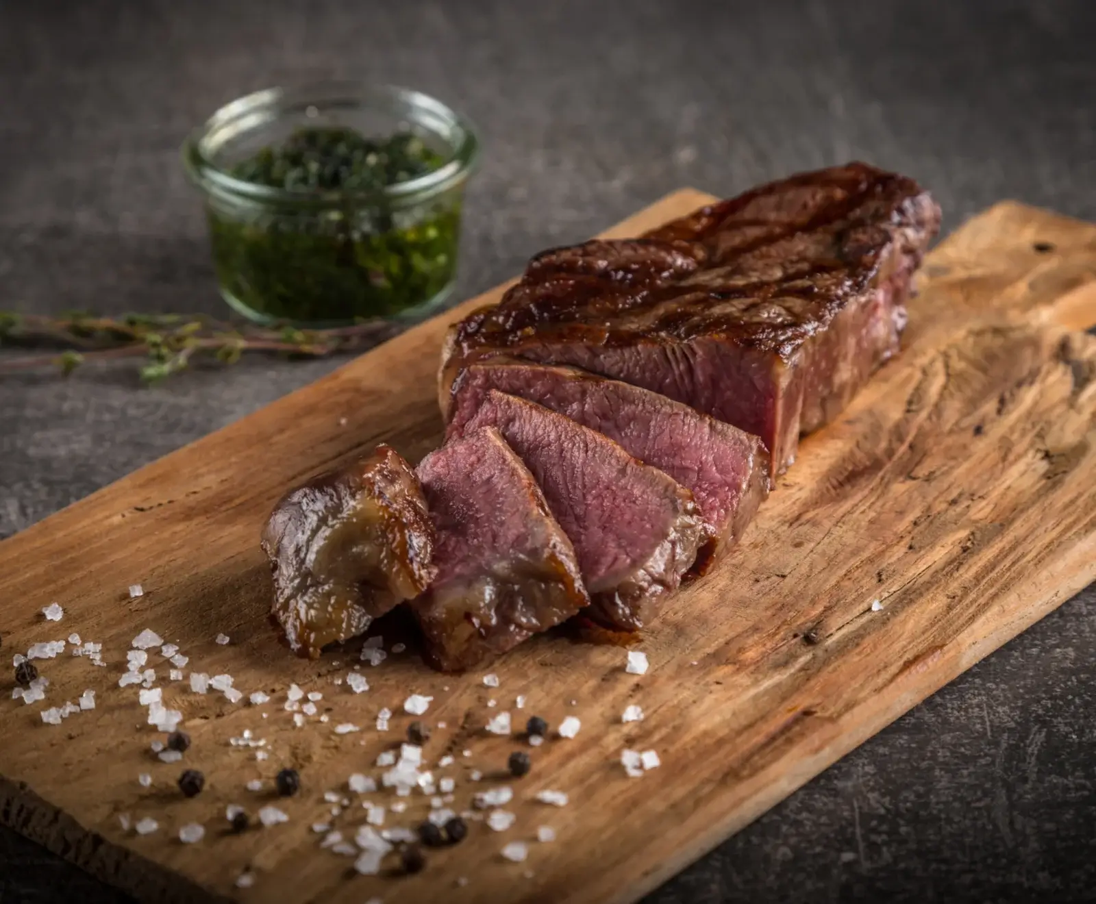 Ein perfekt gebratenes, in Scheiben geschnittenes Steak auf einem Holzbrett, bestreut mit grobem Salz und Pfeffer. Im Hintergrund ein Glas mit grüner Soße.