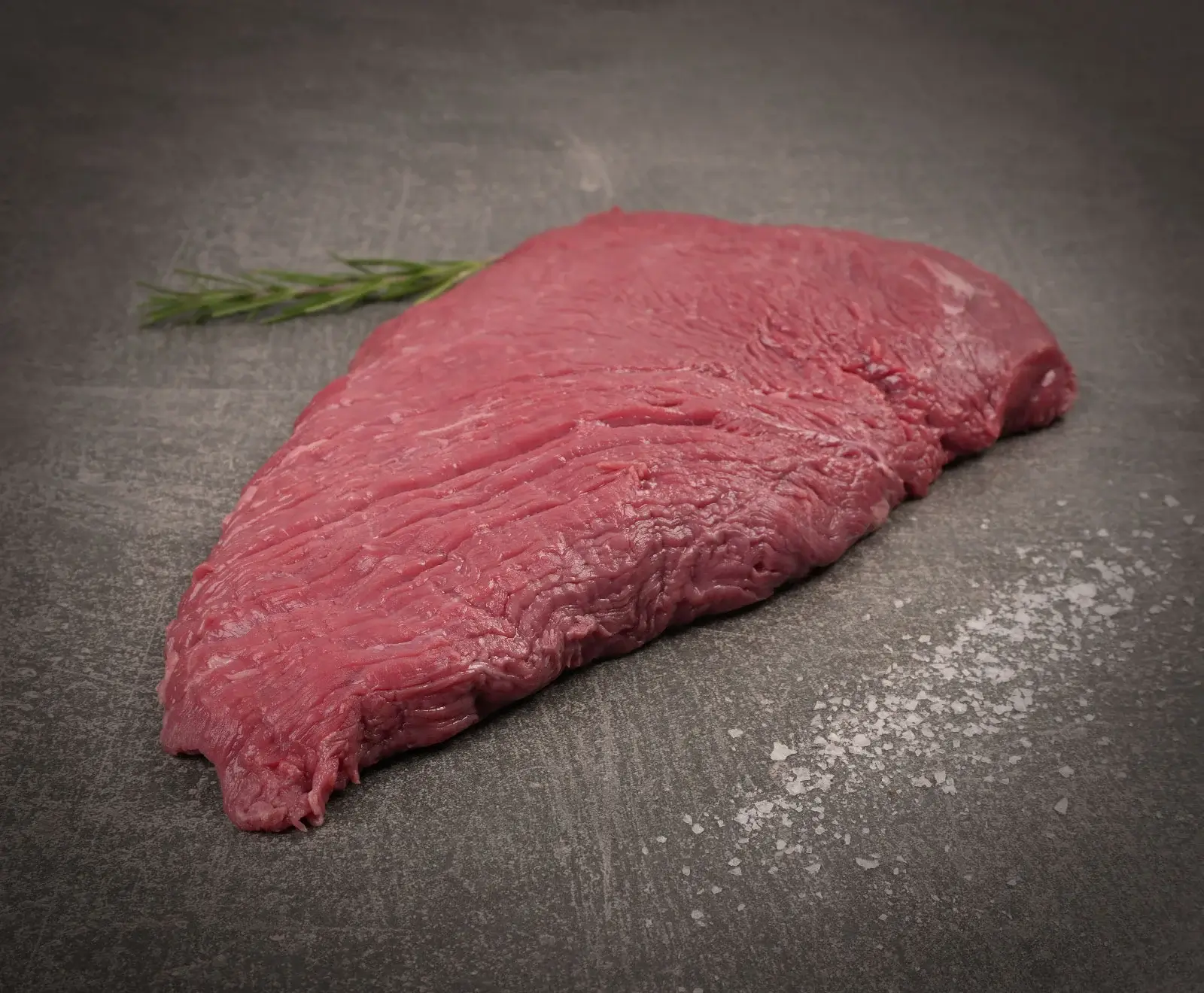 Ein rohes, rotes Stück Rindfleisch liegt auf einer dunklen Steinplatte. Links oben ist ein Rosmarinzweig, rechts unten sind grobe Salzkristalle verstreut.