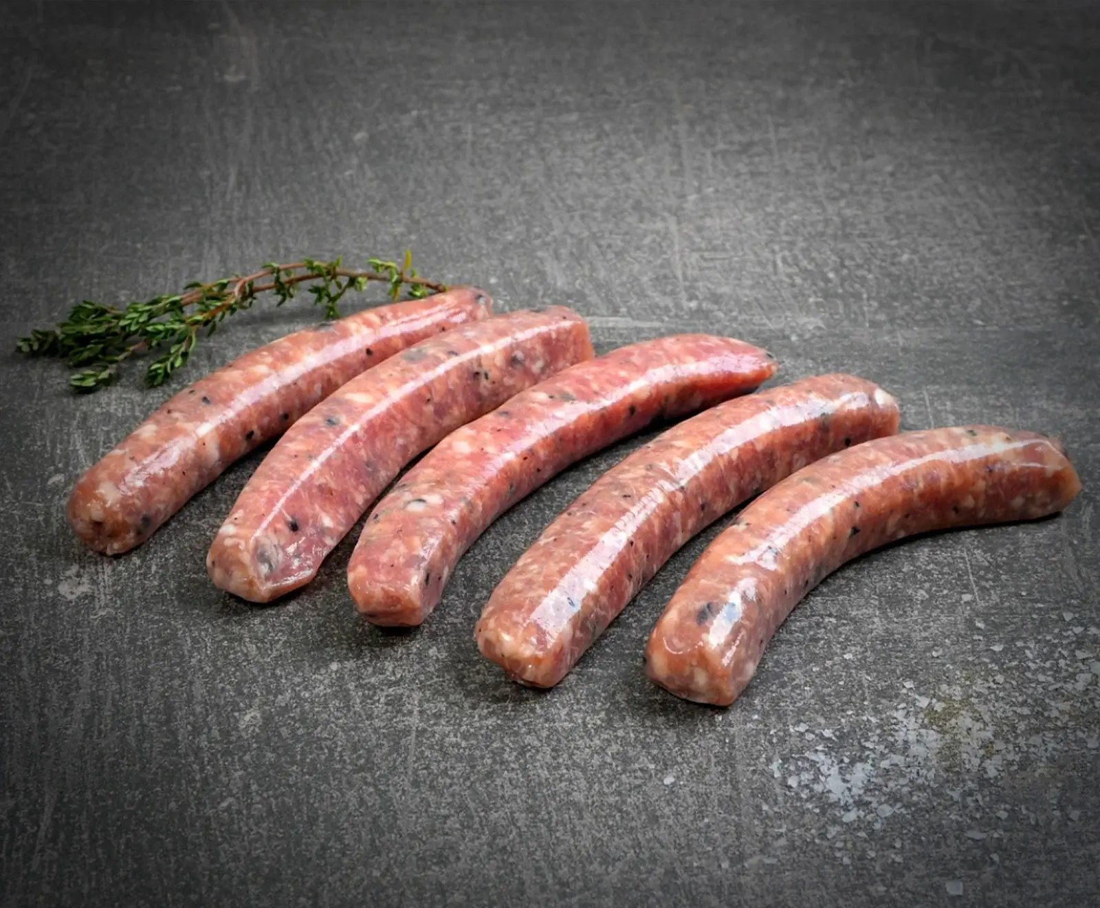 Fünf rohe, hellrosa Würste mit sichtbaren Gewürzsprenkeln liegen leicht gebogen auf einer dunklen, strukturierten Oberfläche. Ein kleiner Thymianzweig ist im oberen linken Bereich zu sehen.