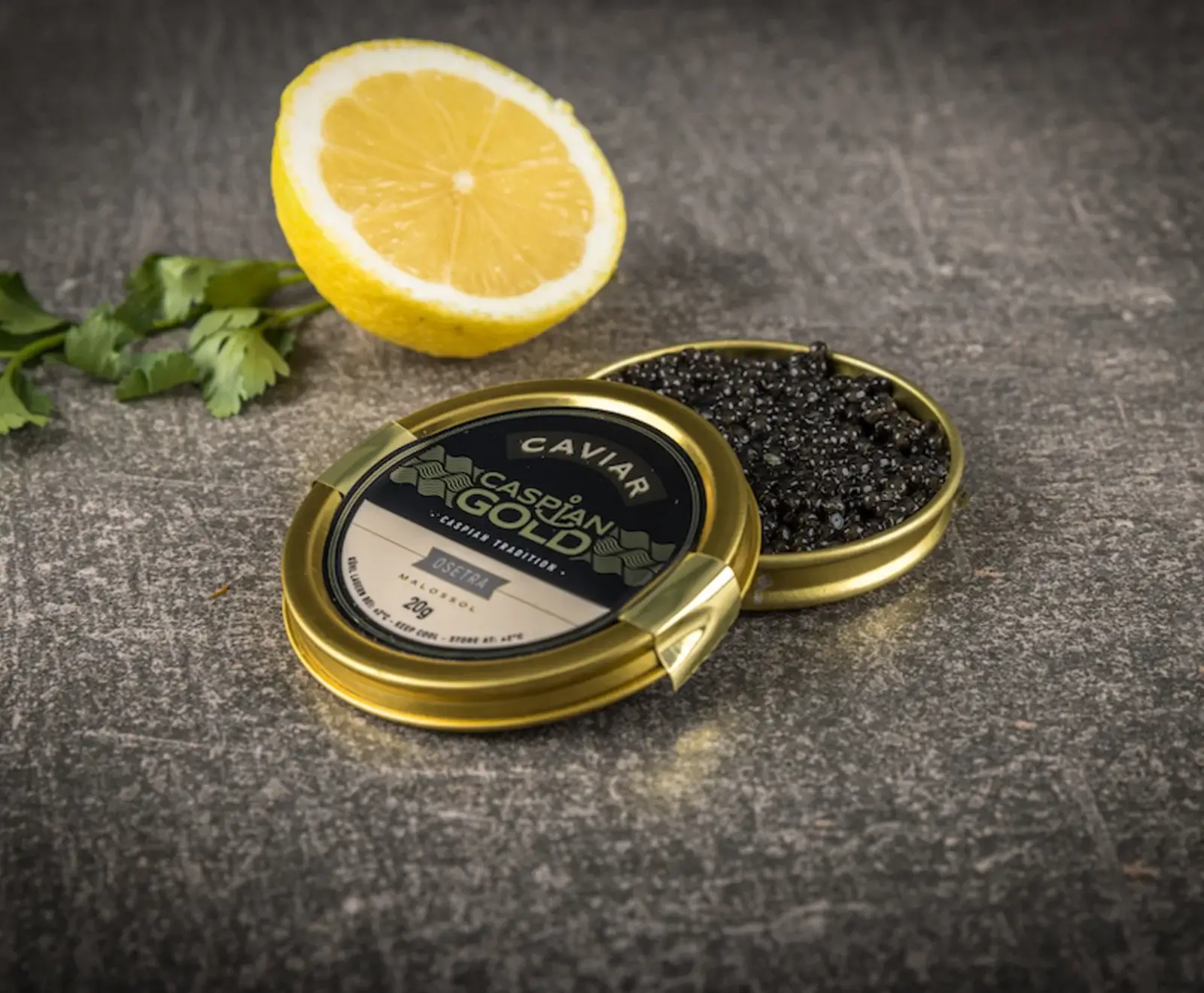 Eine offene goldene Dose "Caspian Gold Osetra" Kaviar mit schwarzem Kaviar, daneben eine halbe Zitrone und Petersilie auf dunklem, strukturiertem Untergrund.