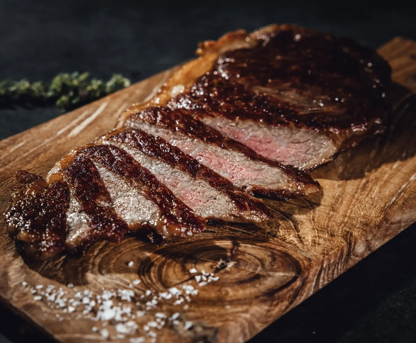 Ein perfekt gebratenes, in Scheiben geschnittenes Steak mit rosa Kern auf einem rustikalen Holzbrett, bestreut mit grobem Meersalz. Im unscharfen Hintergrund sind Kräuter zu sehen.