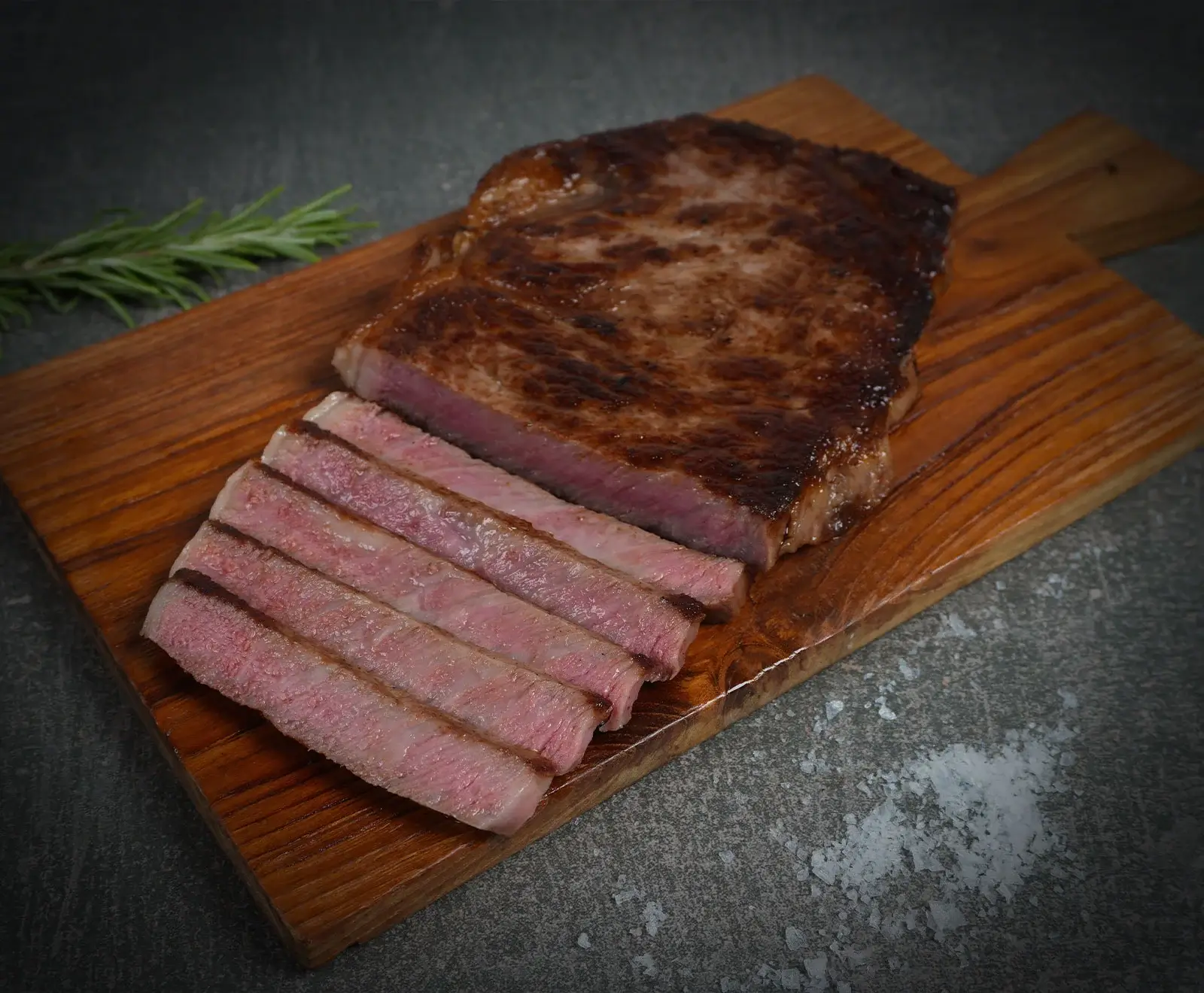 Ein perfekt gebratenes, medium-rare Rindersteak, teilweise in Scheiben geschnitten, liegt auf einem Holzschneidebrett. Daneben ein Rosmarinzweig und grobes Salz auf dunklem Untergrund.