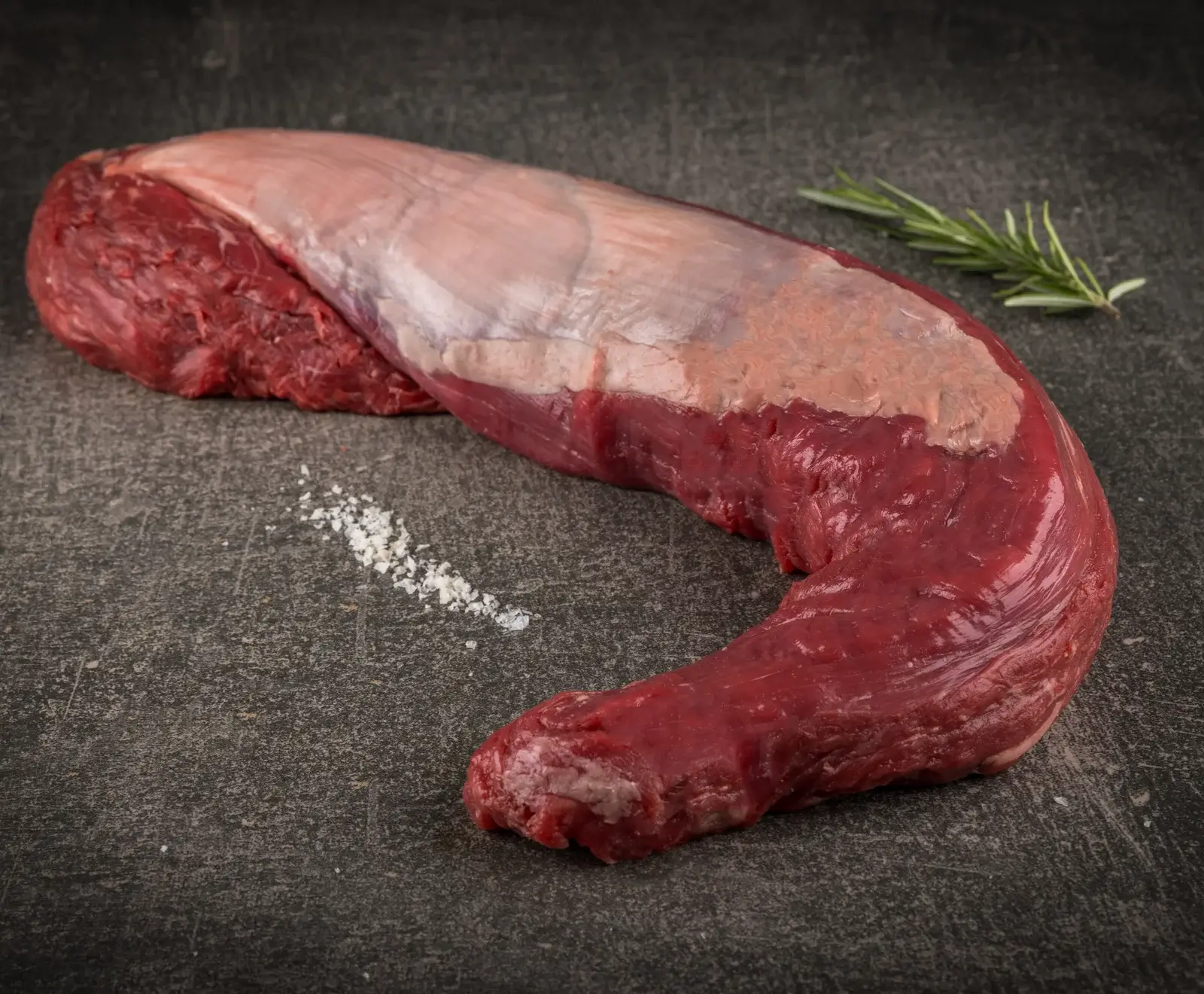 Rohes Rinderfilet mit Silberhaut auf dunklem Untergrund, daneben grobes Salz und ein Rosmarinzweig.