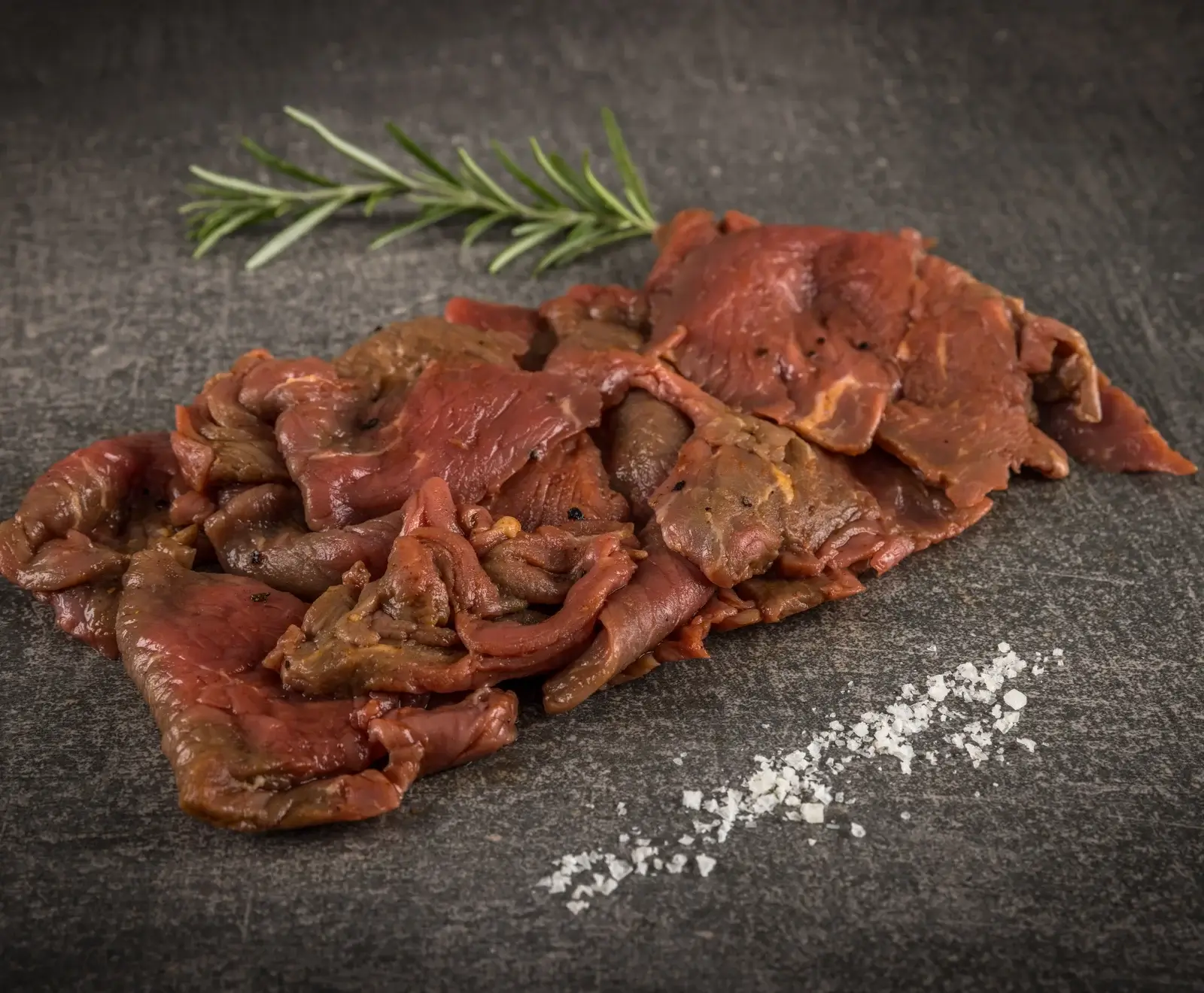 Ein Haufen dünn geschnittenes, mariniertes rotes Fleisch auf einer dunklen Steinplatte, garniert mit einem Rosmarinzweig und grobem Salz.