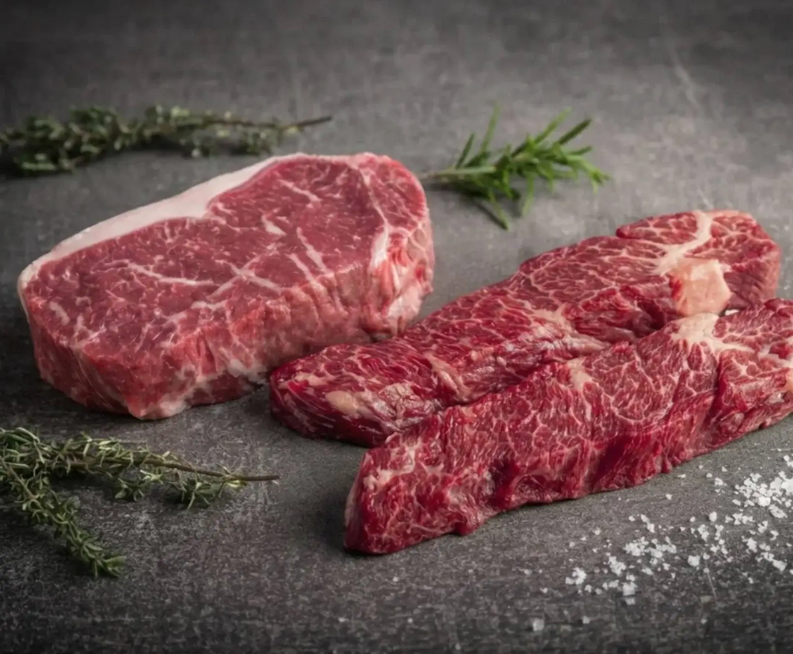 Rohes Rindfleisch: Ein dickes Steak und zwei flachere Stücke mit Marmorierung liegen auf dunklem Untergrund, umgeben von Rosmarin, Thymian und grobem Salz.