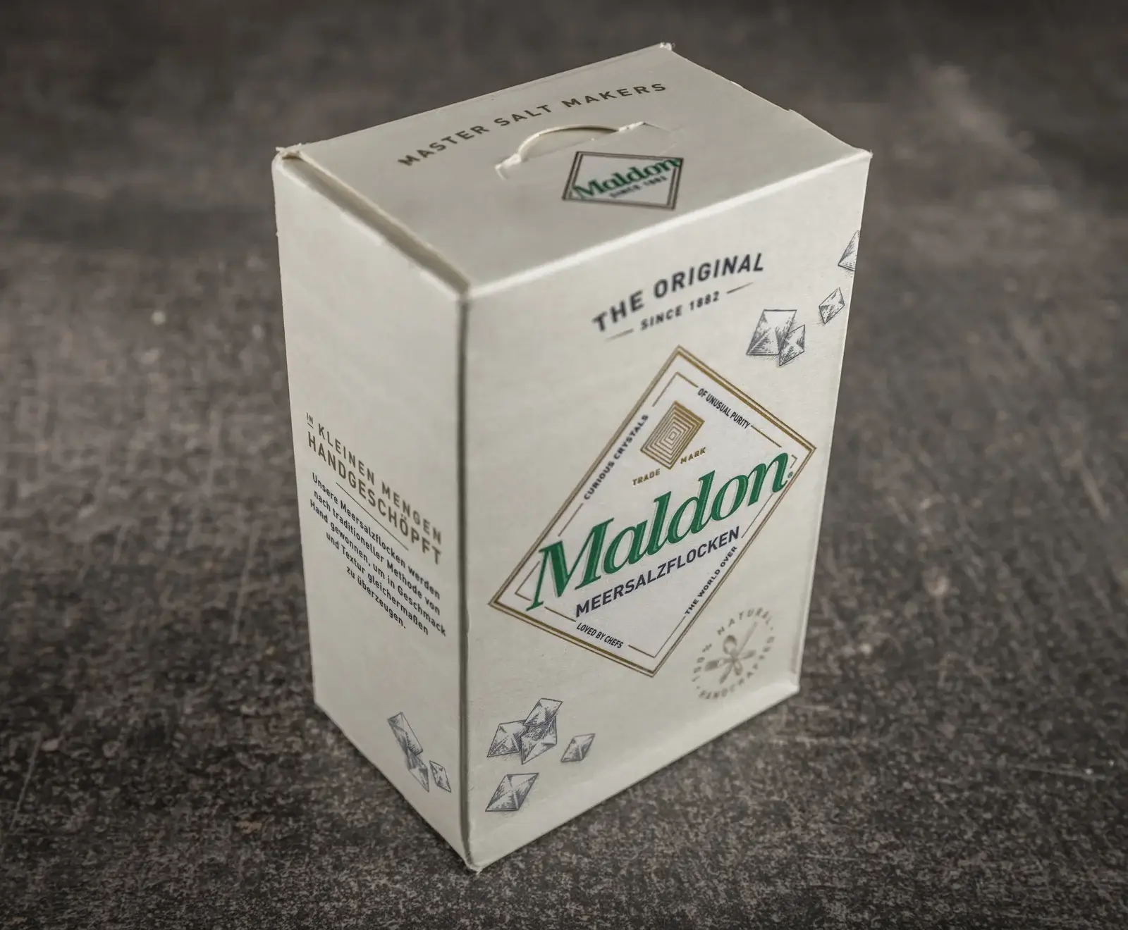Eine weiße Kartonverpackung von Maldon Meersalzflocken, schräg auf dunklem Hintergrund. Das Etikett zeigt den grünen Maldon-Schriftzug und „Meersalzflocken“. Texte wie „THE ORIGINAL SINCE 1882“ und „IN KLEINEN MENGEN HANDGESCHÖPFT“ sind si