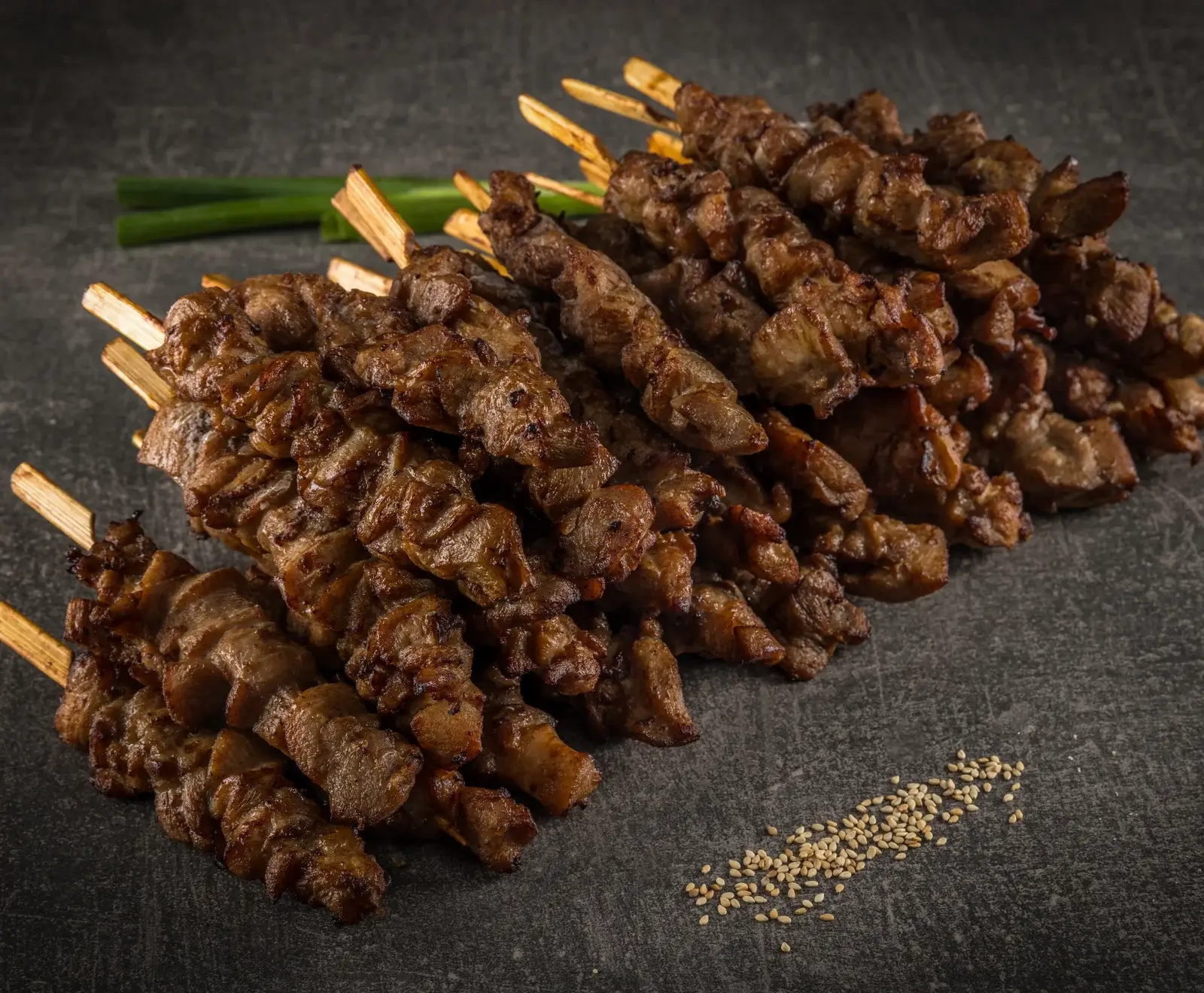 Ein großer Haufen goldbraun gegrillter Fleischspieße auf einem dunklen Untergrund. Im Hintergrund sind grüne Zwiebeln zu sehen, im Vordergrund liegen verstreute Sesamkörner.