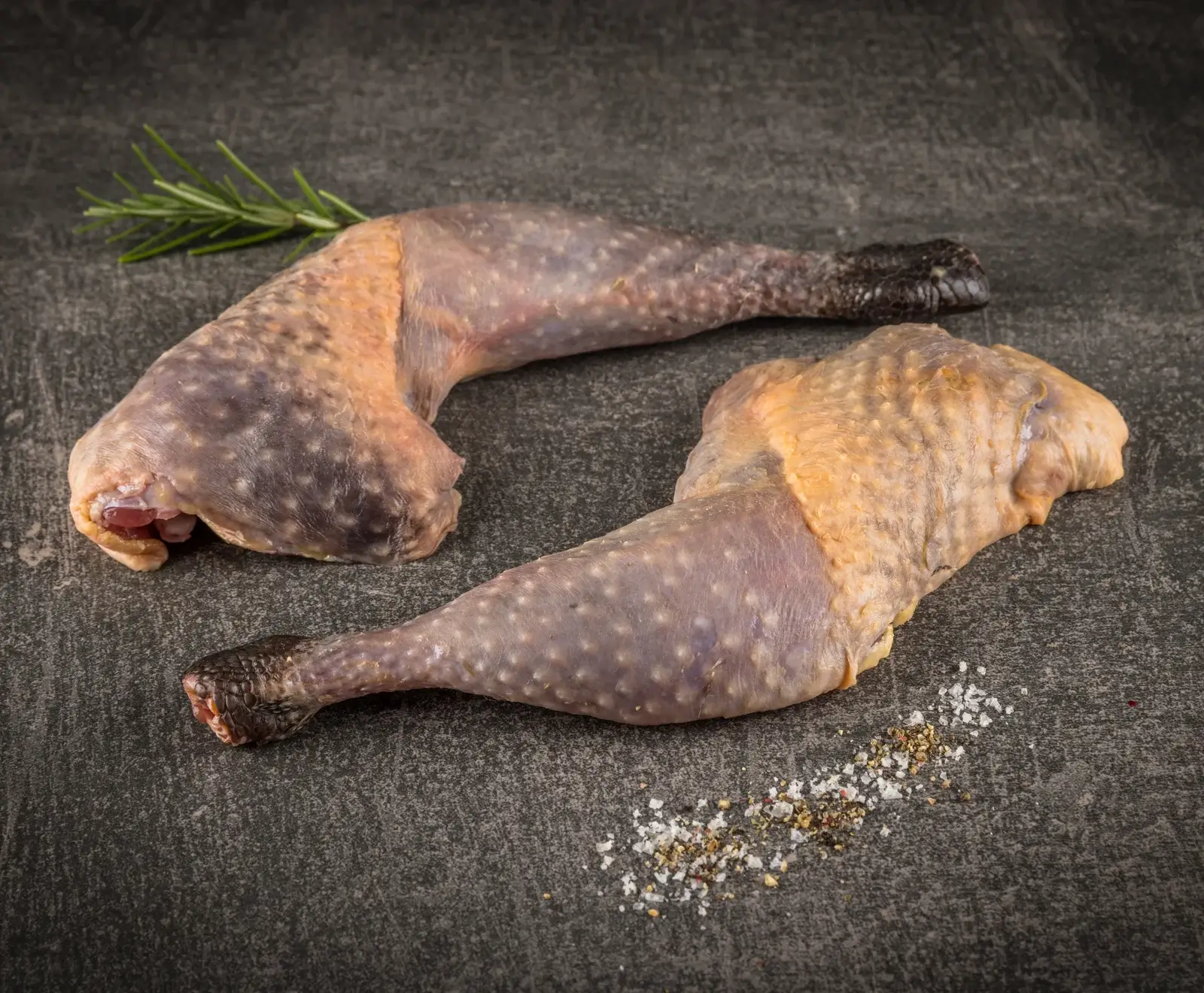 Zwei rohe Wildgeflügelkeulen mit dunkler, teils gelblicher Haut, liegen auf einem dunklen Untergrund. Eine Keule ist mit Rosmarin, die andere mit Salz und Pfeffer garniert.