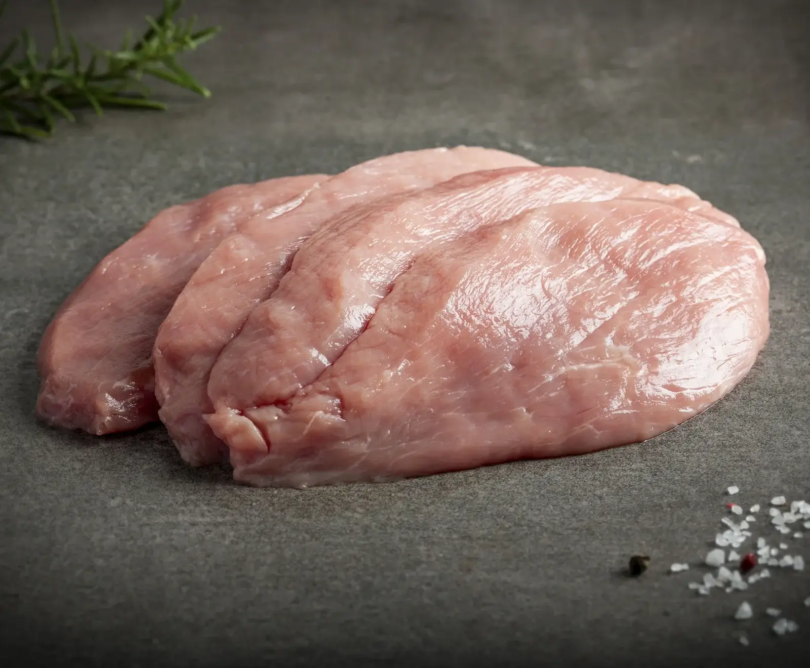 Rohes Hühnerfleisch auf dunklem Hintergrund mit Kräutern und Salz daneben.