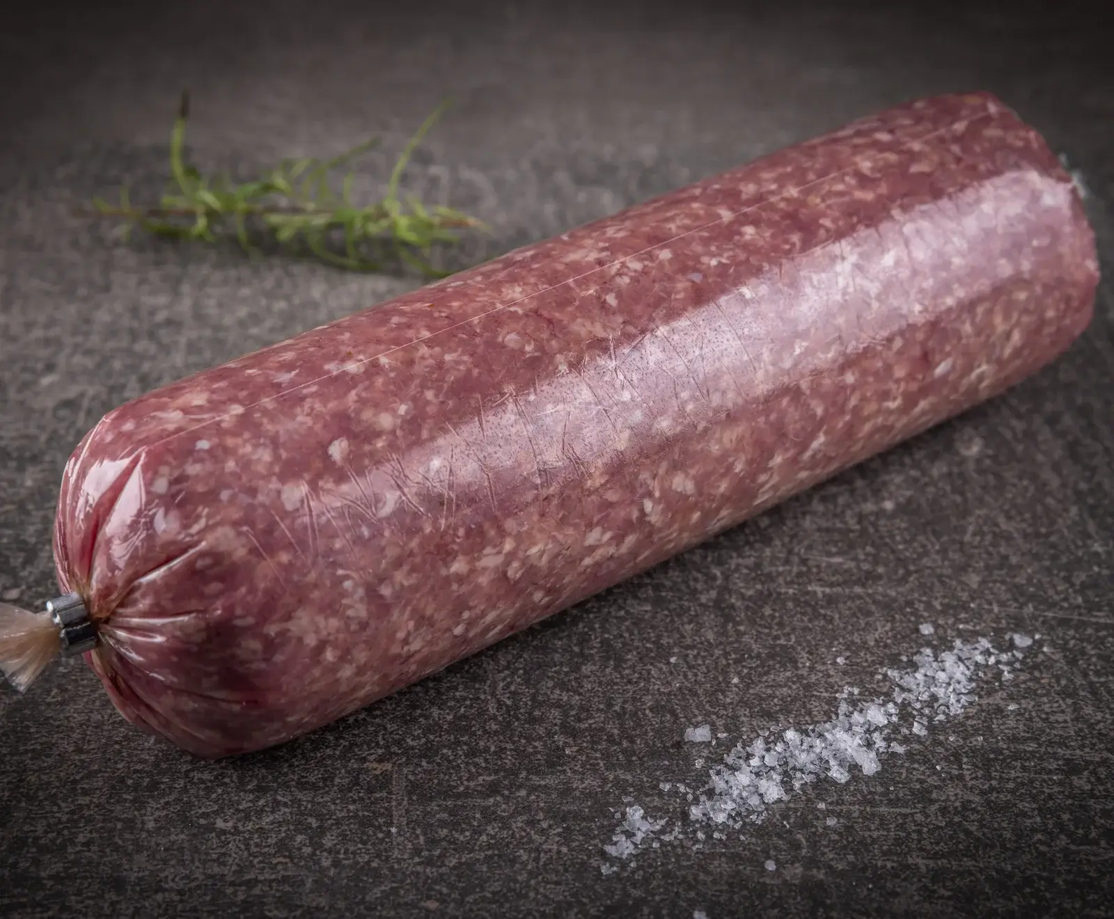 Eine lange, zylindrische Rolle rohes Hackfleisch (Mettwurst) in transparenter Folie, auf dunklem Untergrund. Im Hintergrund Rosmarin, im Vordergrund grobes Salz.