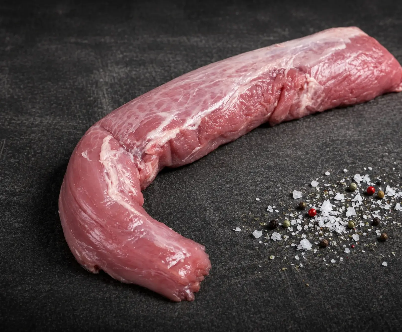 Ein rohes, leicht gekrümmtes Schweinefilet liegt auf einer dunklen, strukturierten Oberfläche. Daneben sind grobes Meersalz und bunte Pfefferkörner verstreut.