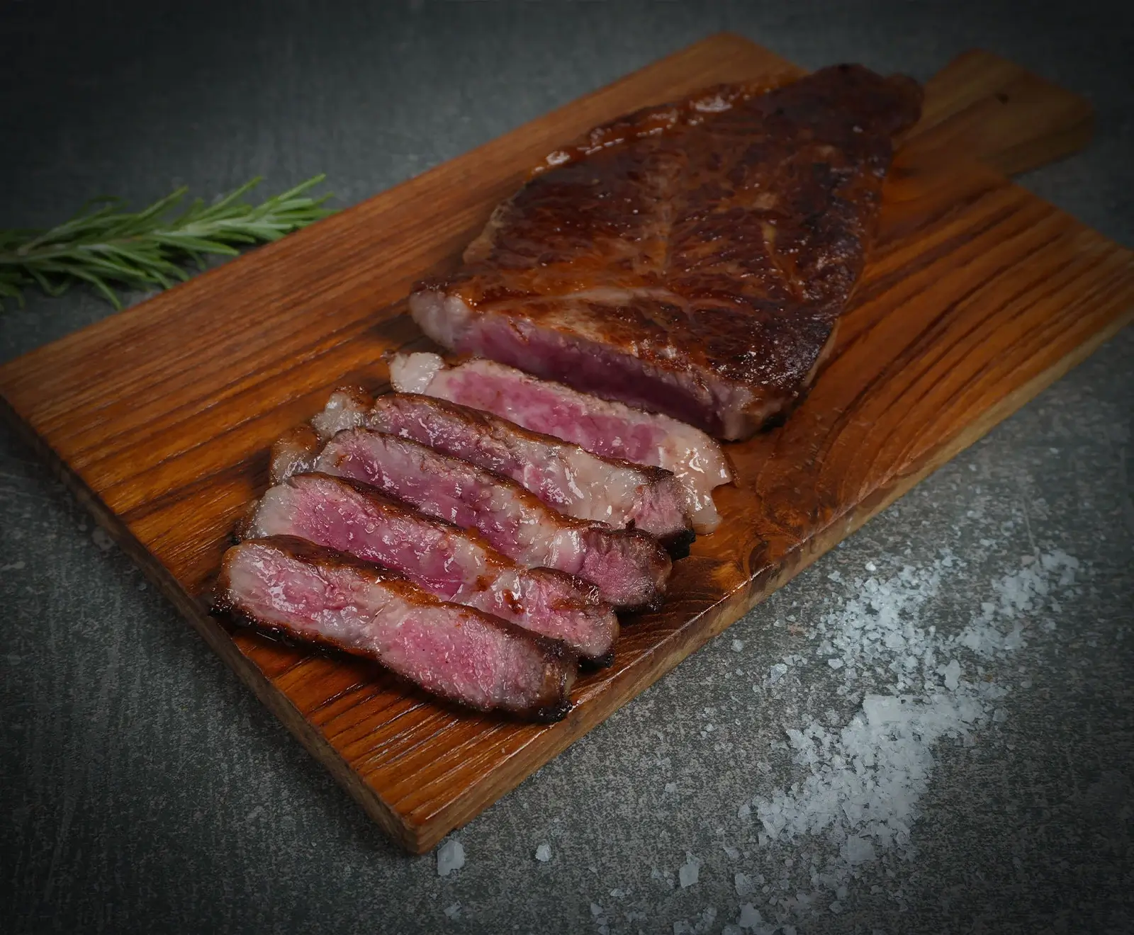 Ein saftiges, medium-rare gebratenes Steak, teilweise in Scheiben geschnitten, liegt auf einem Holzschneidebrett. Daneben sind ein Rosmarinzweig und grobes Meersalz auf dunklem Untergrund zu sehen.