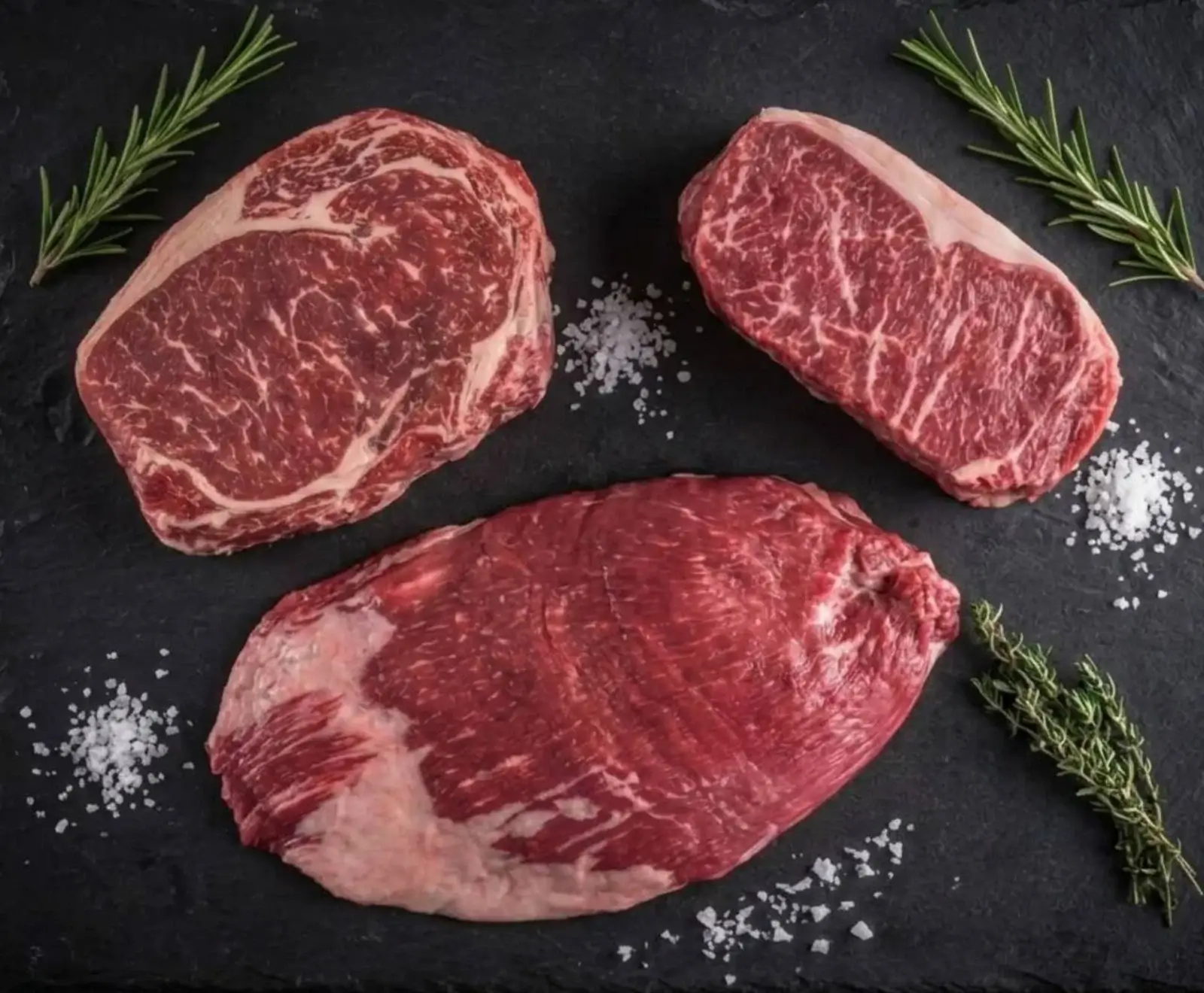 Drei rohe Rindersteaks (Ribeye, Striploin, Flanksteak) auf dunklem Schiefer, garniert mit Rosmarin, Thymian und grobem Meersalz. Die Steaks zeigen eine schöne Marmorierung.