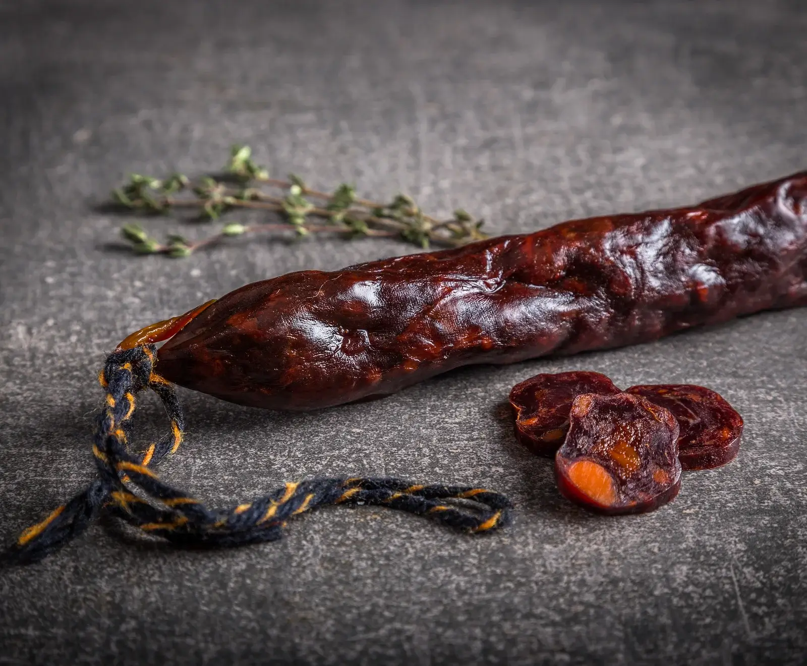 Eine dunkelrote, glänzende Wurst mit blau-gelbem Faden, daneben drei Scheiben, auf dunklem Steinhintergrund. Im unscharfen Hintergrund liegen Thymianzweige.