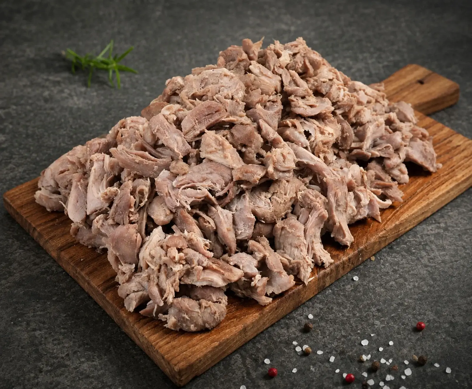Ein großer Haufen zerkleinertes, gekochtes Fleisch auf einem Holzschneidebrett. Im Hintergrund ein Rosmarinzweig, im Vordergrund grobes Salz und Pfefferkörner auf dunklem Untergrund.