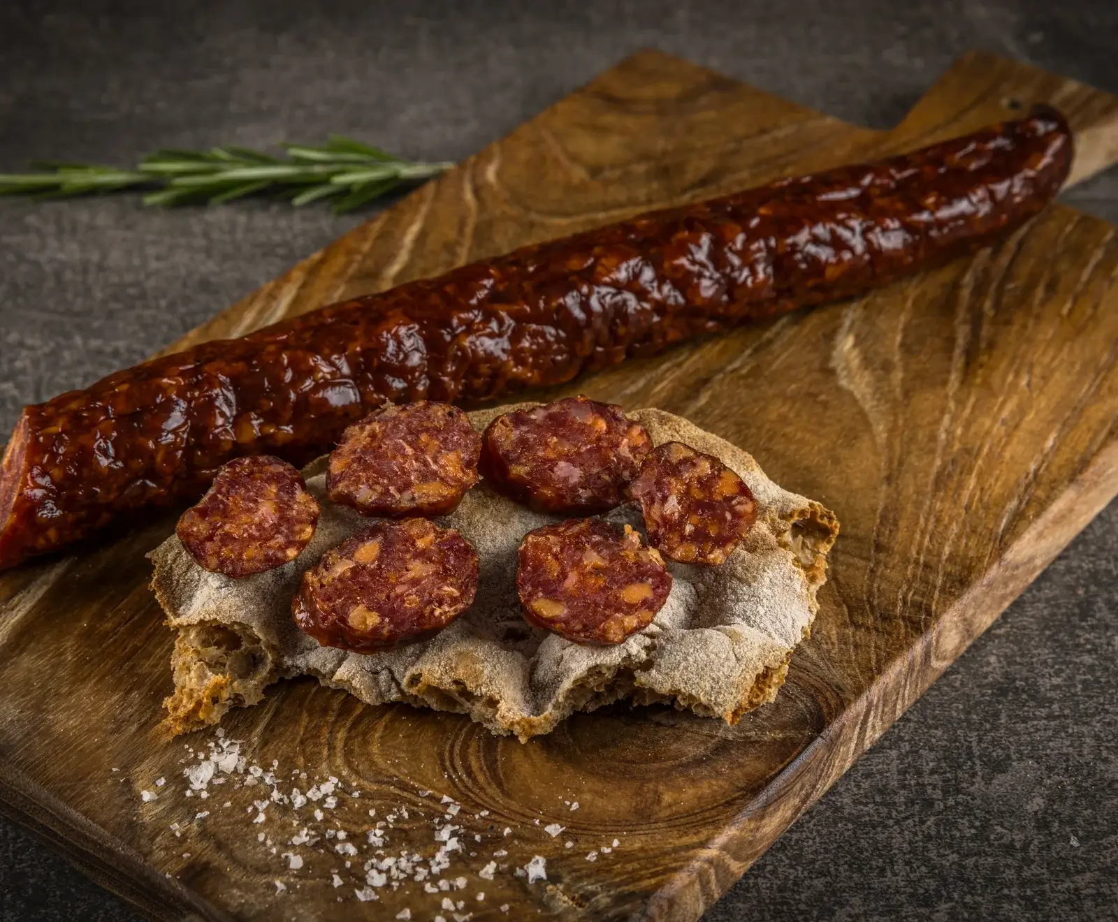 Eine lange, dunkelrote Wurst liegt auf einem Holzbrett. Daneben ein Stück rustikales Brot mit mehreren Scheiben der Wurst, bestreut mit grobem Meersalz. Im Hintergrund ein Rosmarinzweig.