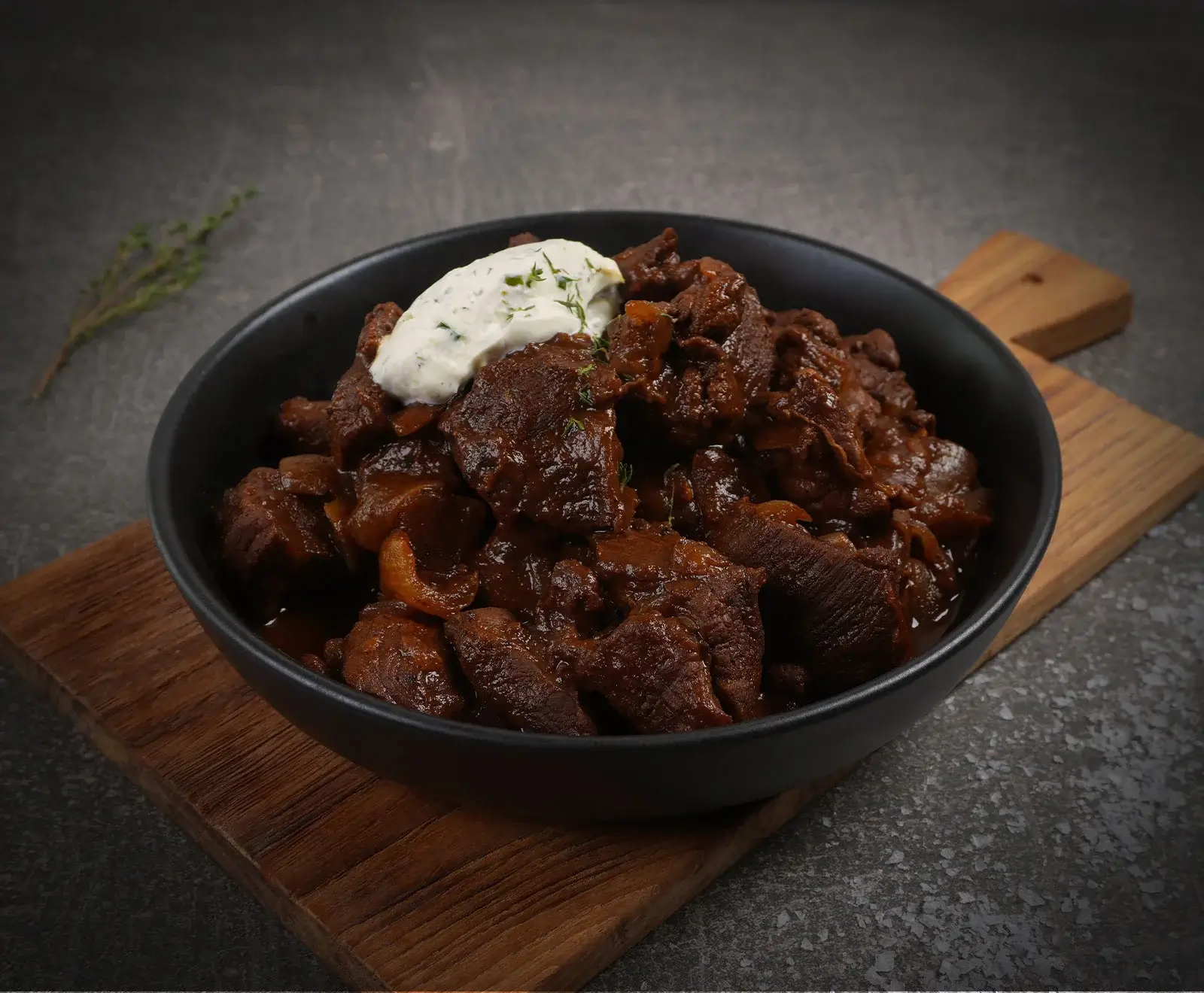 Eine schwarze Schüssel gefüllt mit dunklem Rindereintopf oder Gulasch, garniert mit einem Klecks Sauerrahm und frischem Thymian, steht auf einem Holzbrett vor einem dunklen Hintergrund.