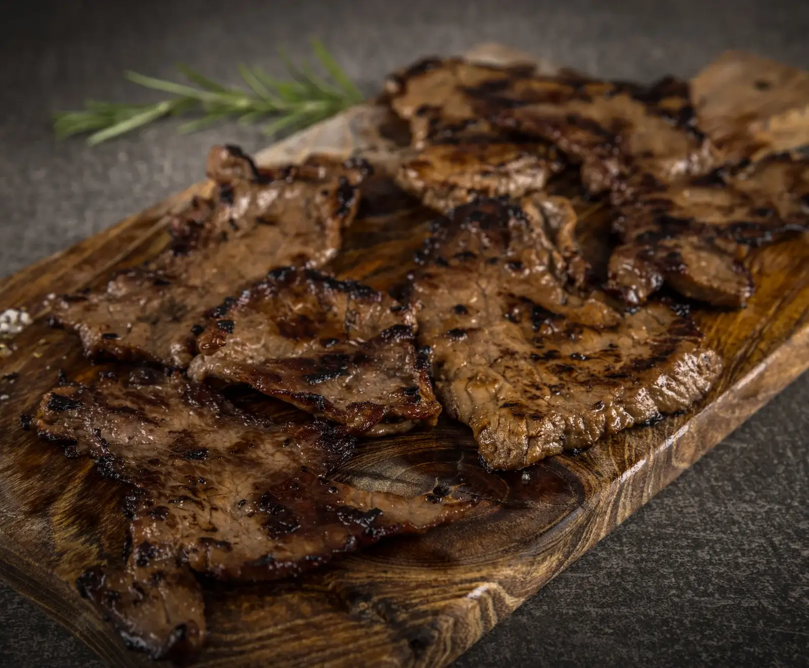 Mehrere dünn geschnittene, gegrillte Fleischstücke mit deutlichen Grillspuren liegen auf einem dunklen Holzbrett. Im unscharfen Hintergrund ist ein Rosmarinzweig zu sehen.
