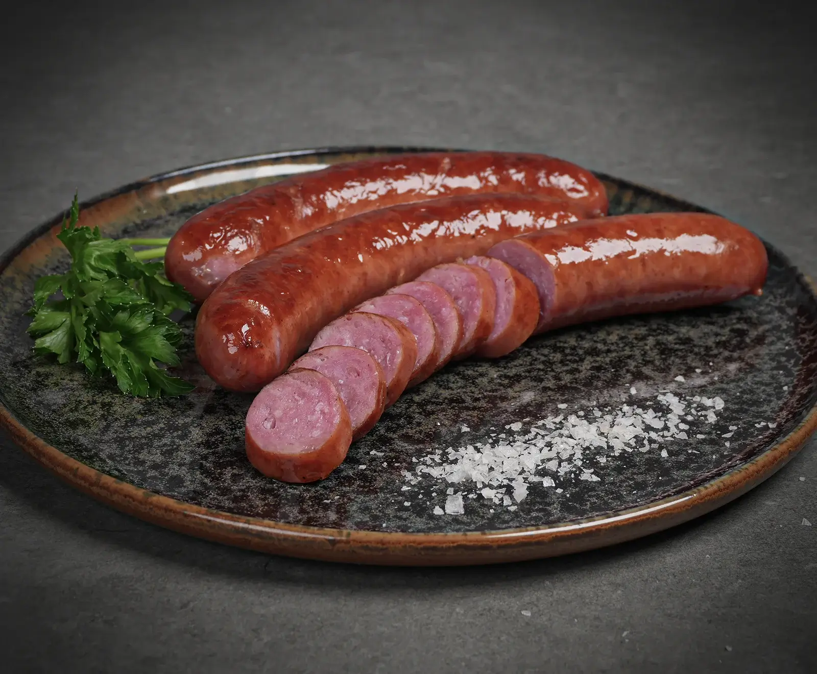 Drei glänzende, rotbraune Würste auf einem dunklen Teller. Eine Wurst ist in Scheiben geschnitten und zeigt ein rosa Inneres. Dazu Petersilie und grobes Salz.